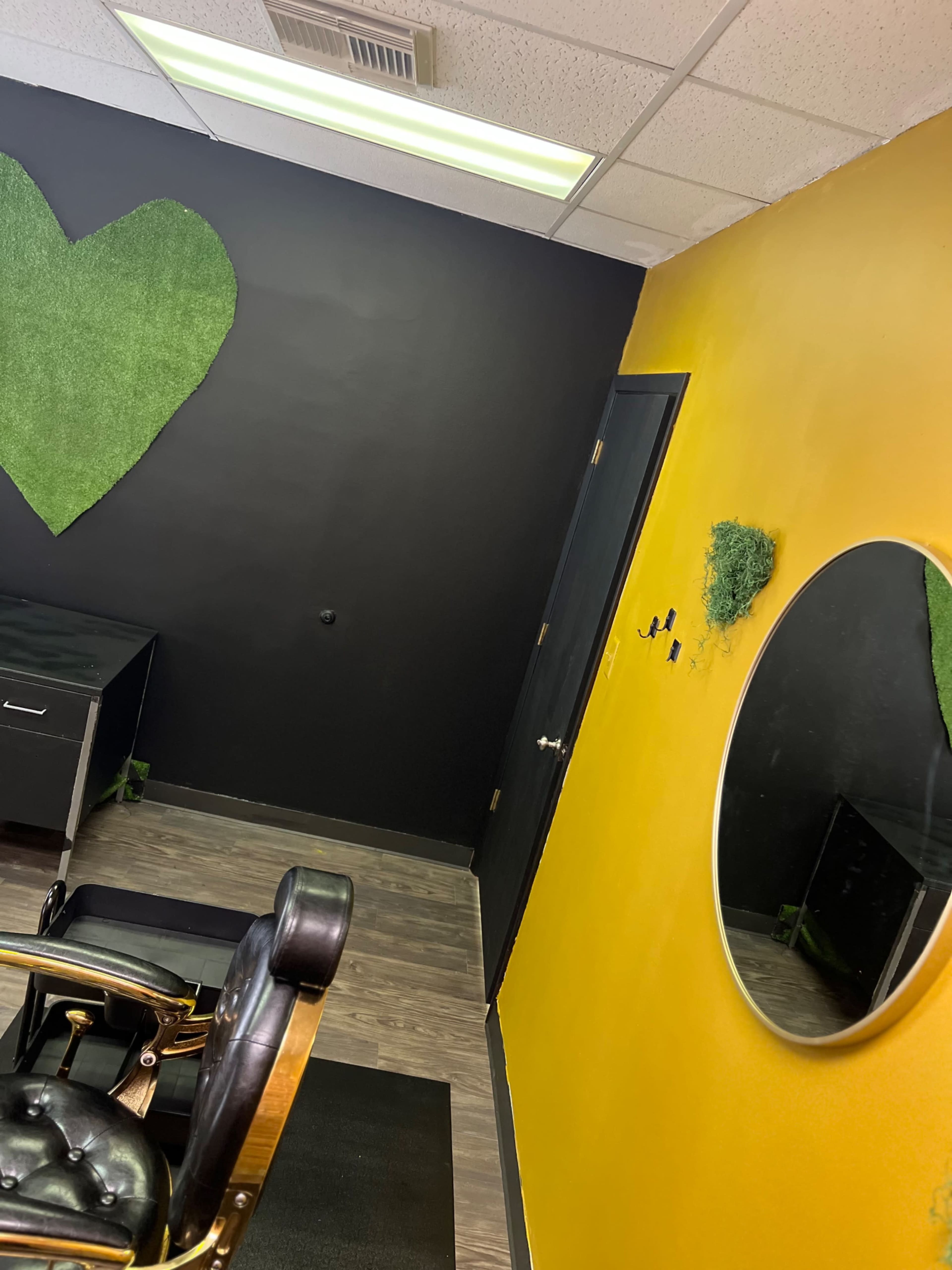 The image shows a salon interior featuring a black wall adorned with artificial greenery and a yellow wall with a circular mirror.