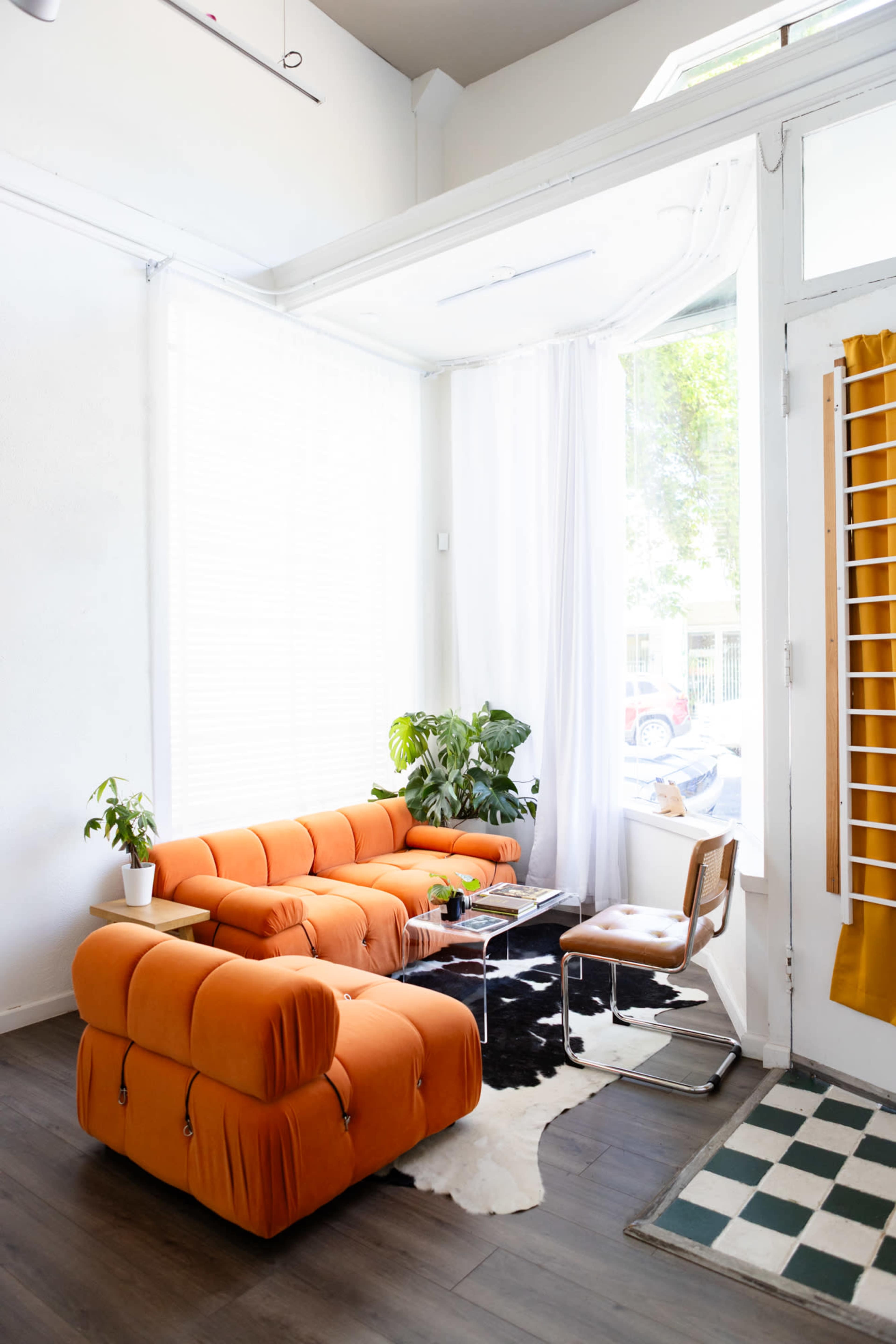 The image shows a modern living room with a bright orange sofa, a small plant on a glass coffee table, and large windows that let in natural light.