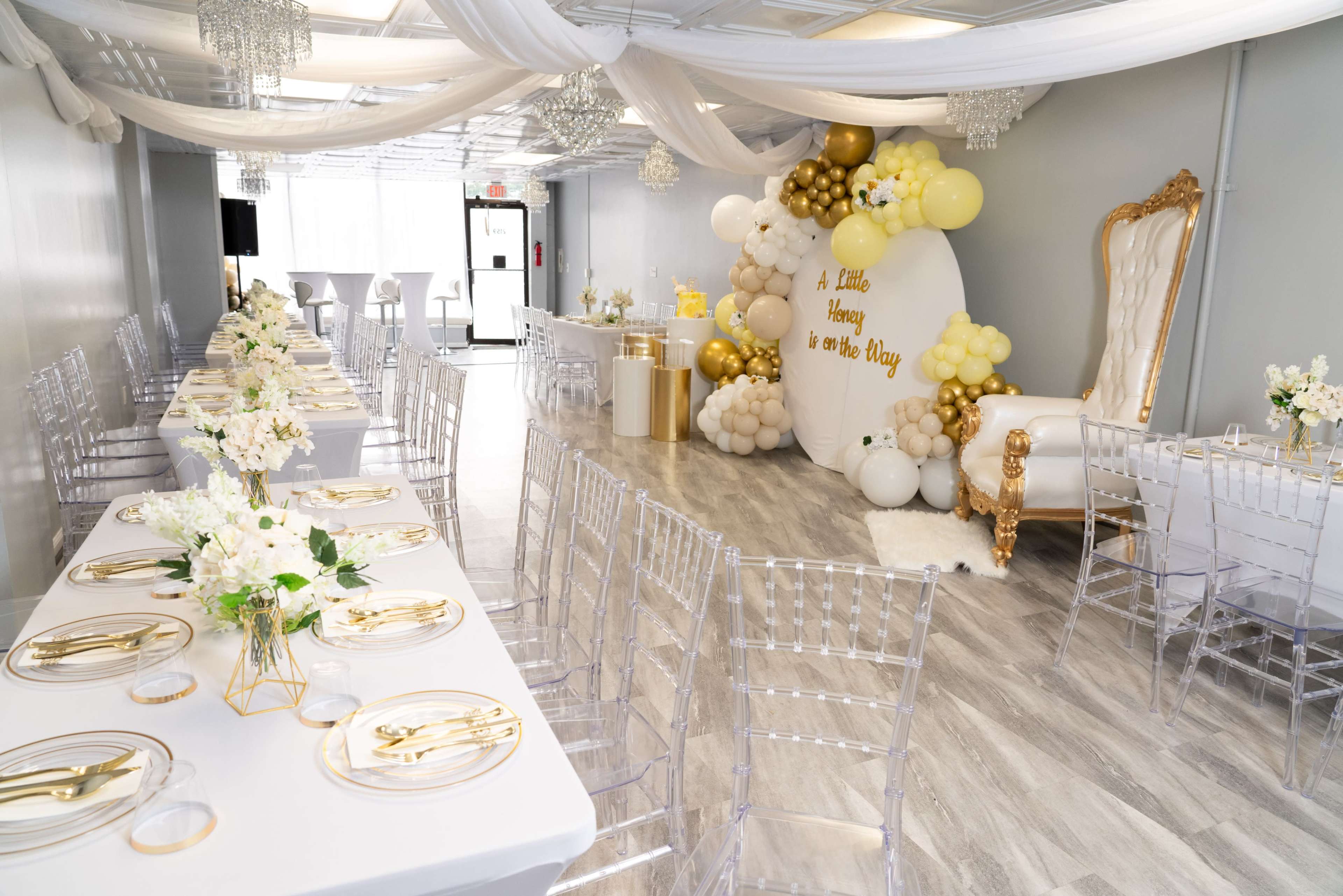 An elegantly decorated event space featuring tables set with gold-accented tableware, a backdrop of balloons, and a large white throne chair.