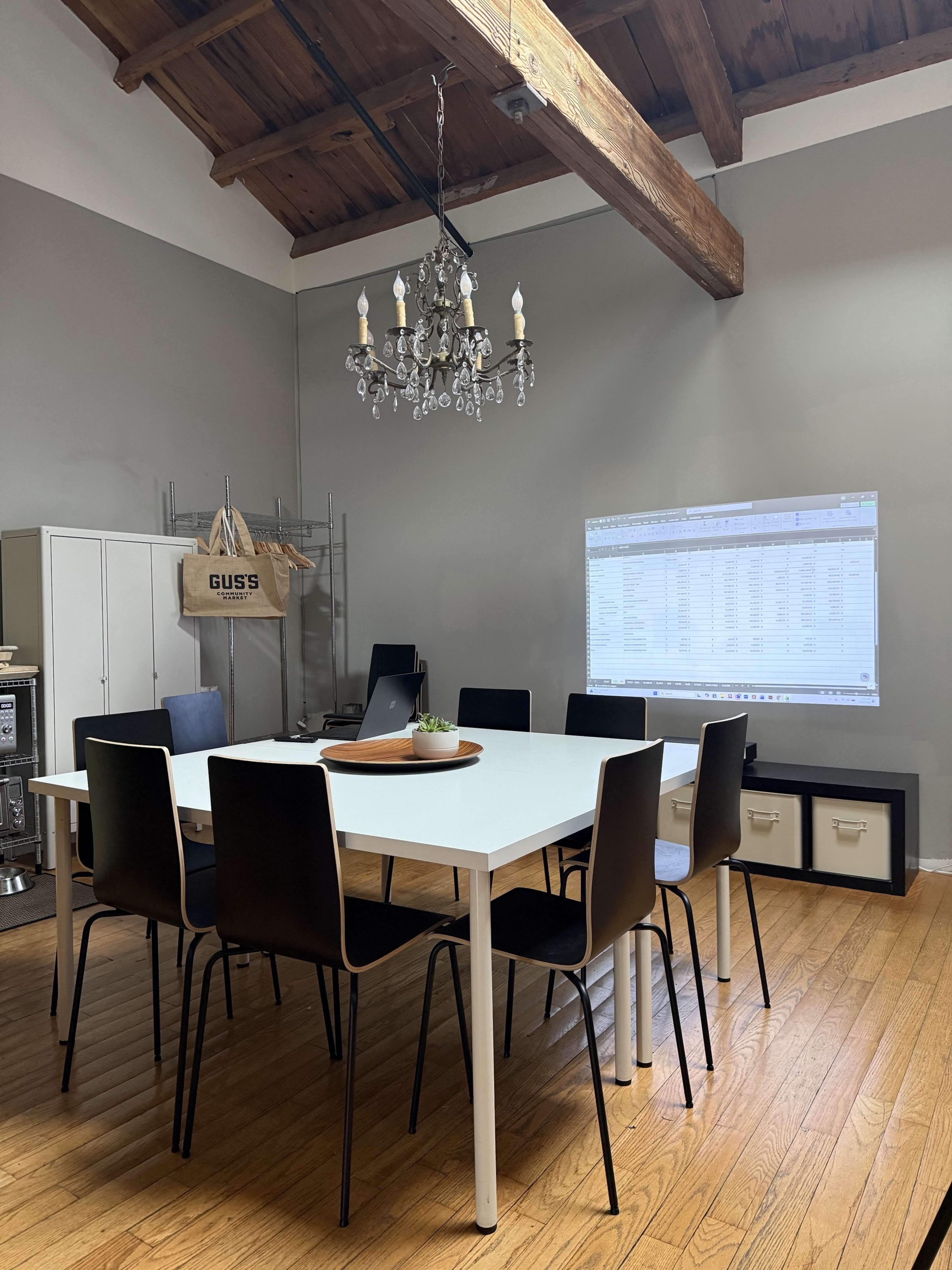The image shows a conference room with a large white table surrounded by black chairs, a chandelier overhead, and a projector displaying a spreadsheet on the wall.