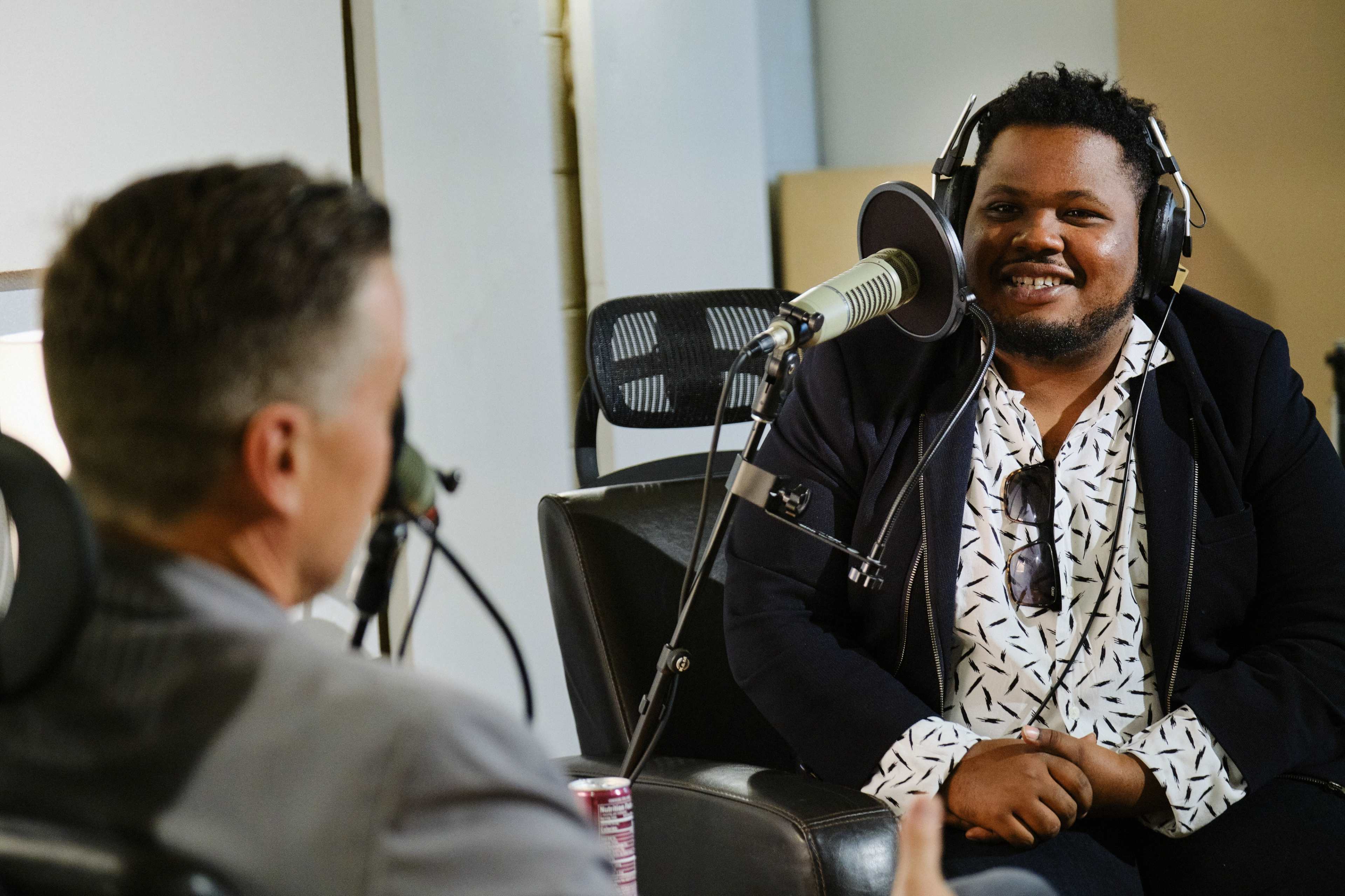 Two men sit across from each other in a podcast studio, with one speaking into a microphone while the other listens.