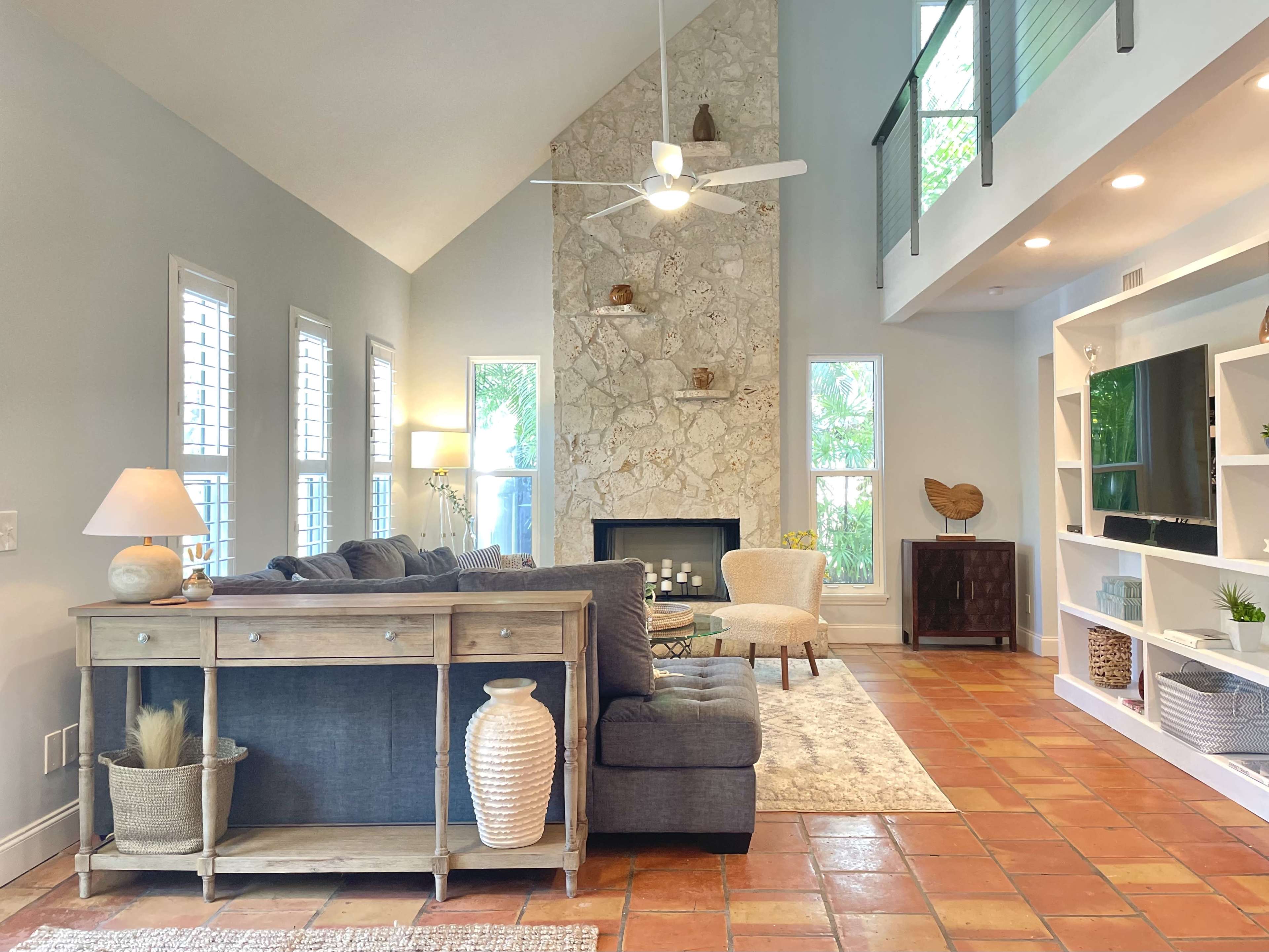 A spacious living room features a gray sofa, a wooden console table, and a stone fireplace with a tall ceiling, illuminated by natural light.