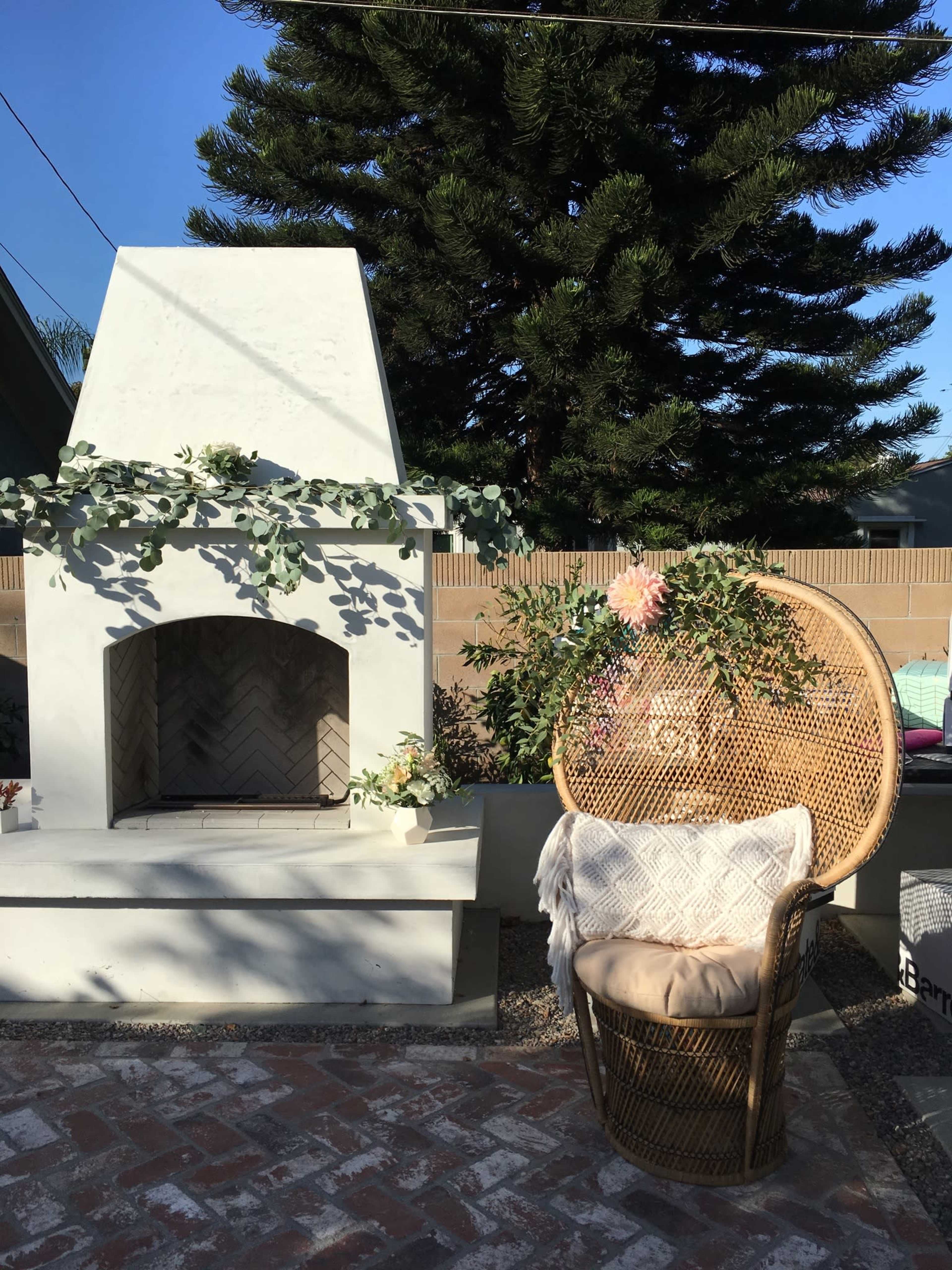 The image shows a cozy outdoor space featuring a white fireplace adorned with greenery, a rattan chair with a cushion, and a tree in the background.
