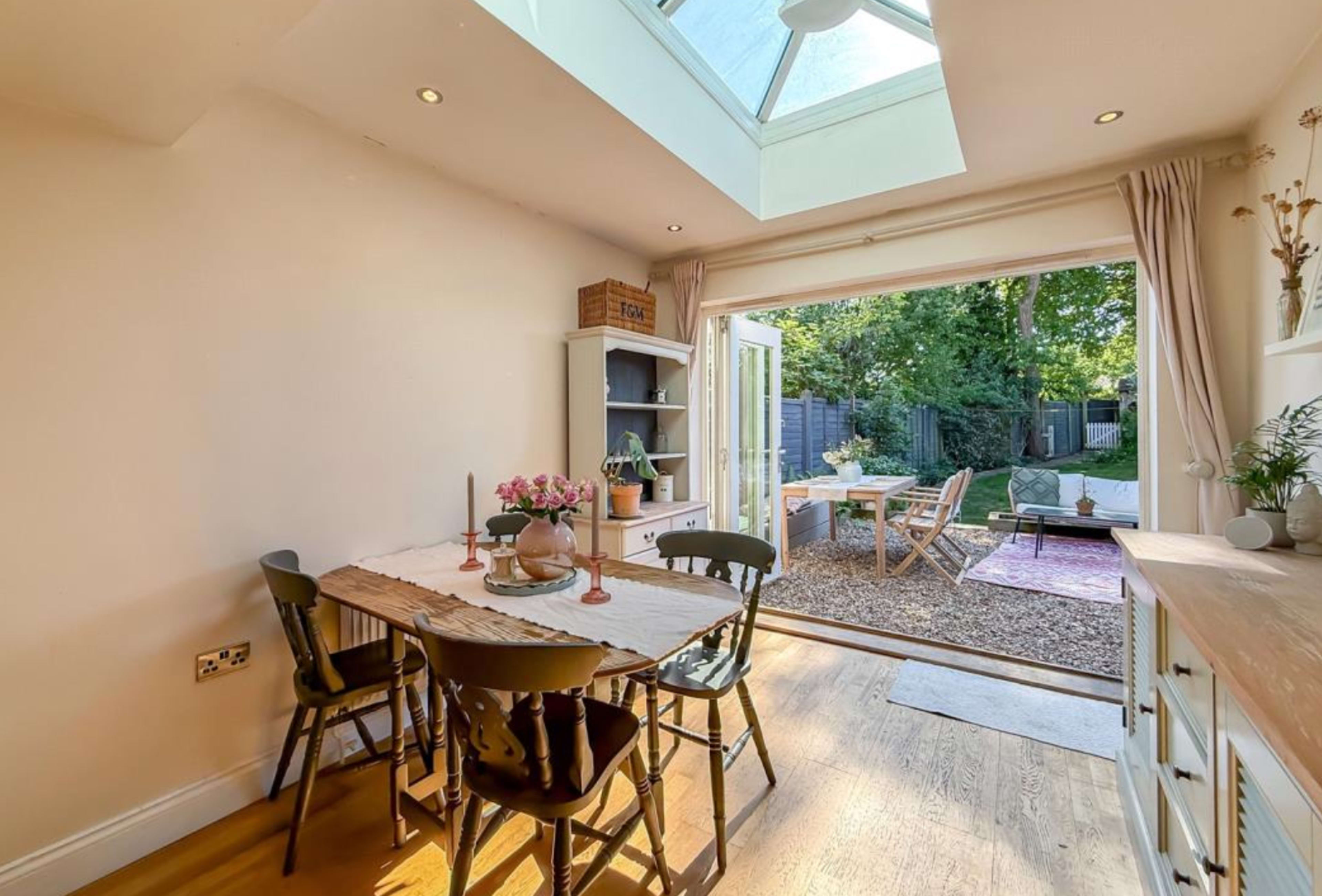 A dining area with a wooden table and chairs opens to a backyard through a glass door, illuminated by natural light from a skylight.
