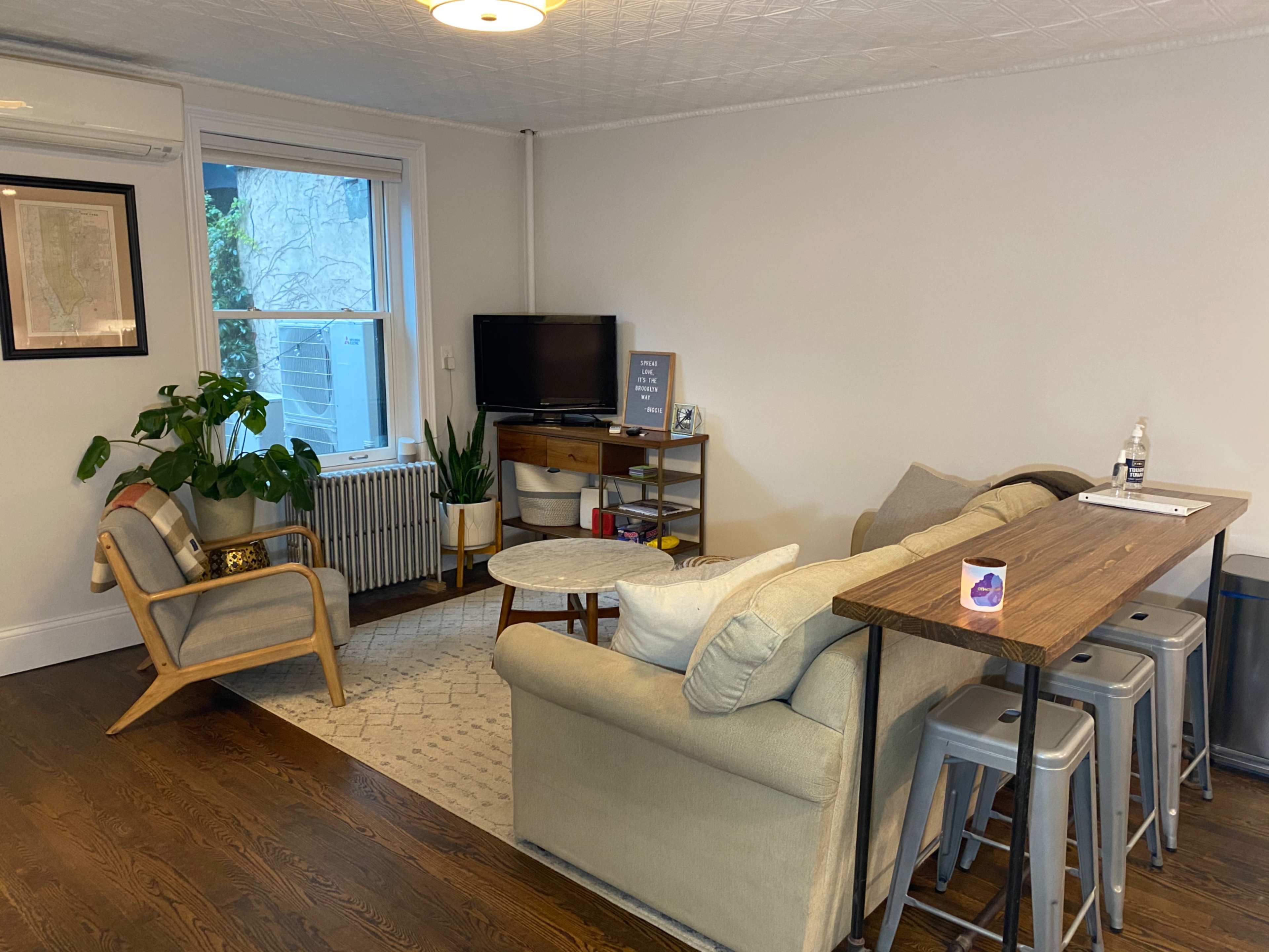 The image shows a modern living room with a sofa, two chairs, a coffee table, a television, and a bar-style dining area with stools.