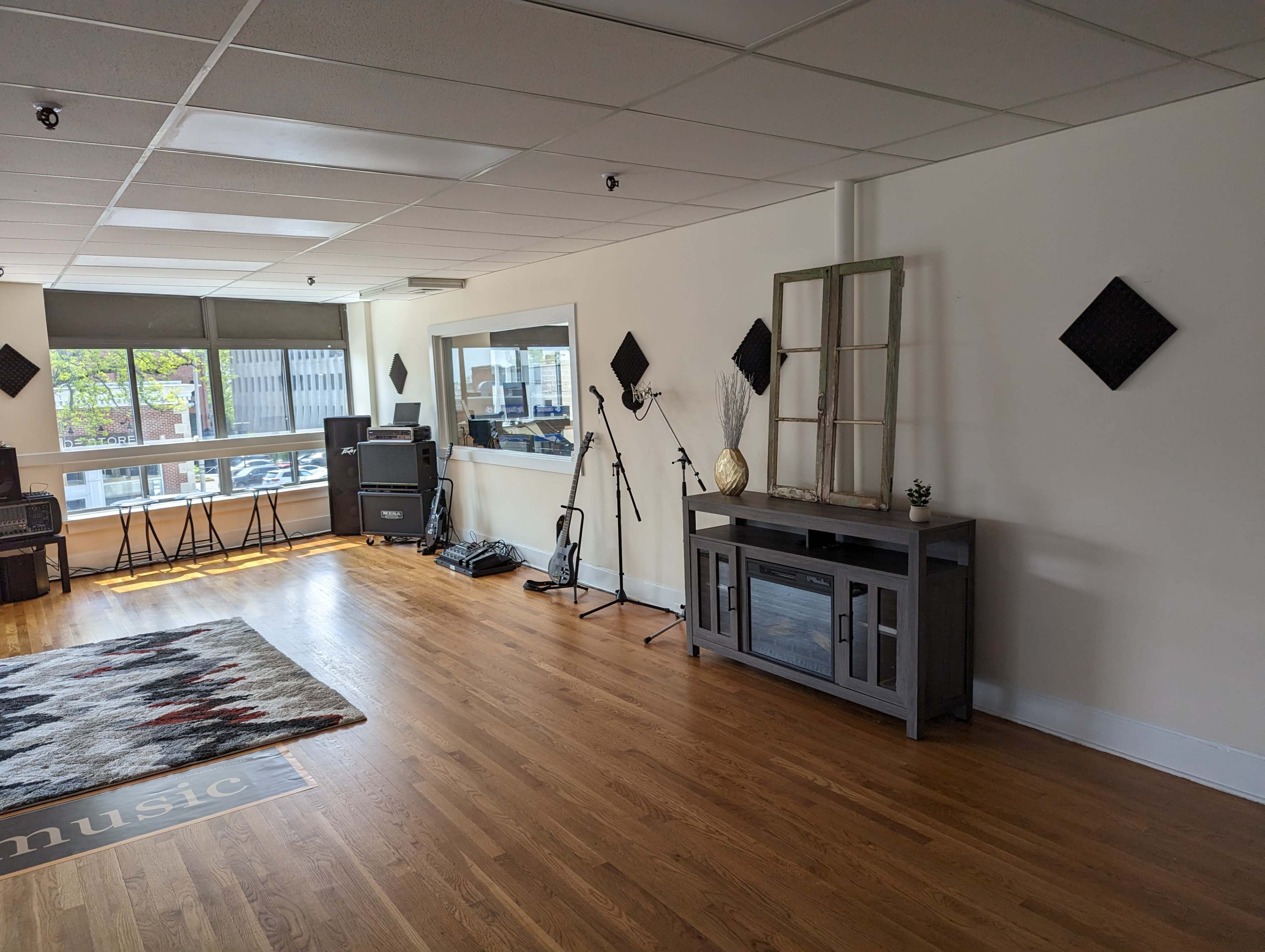 A spacious interior with wooden floors, featuring a music setup against one wall, a decorative cabinet, and large windows letting in natural light.