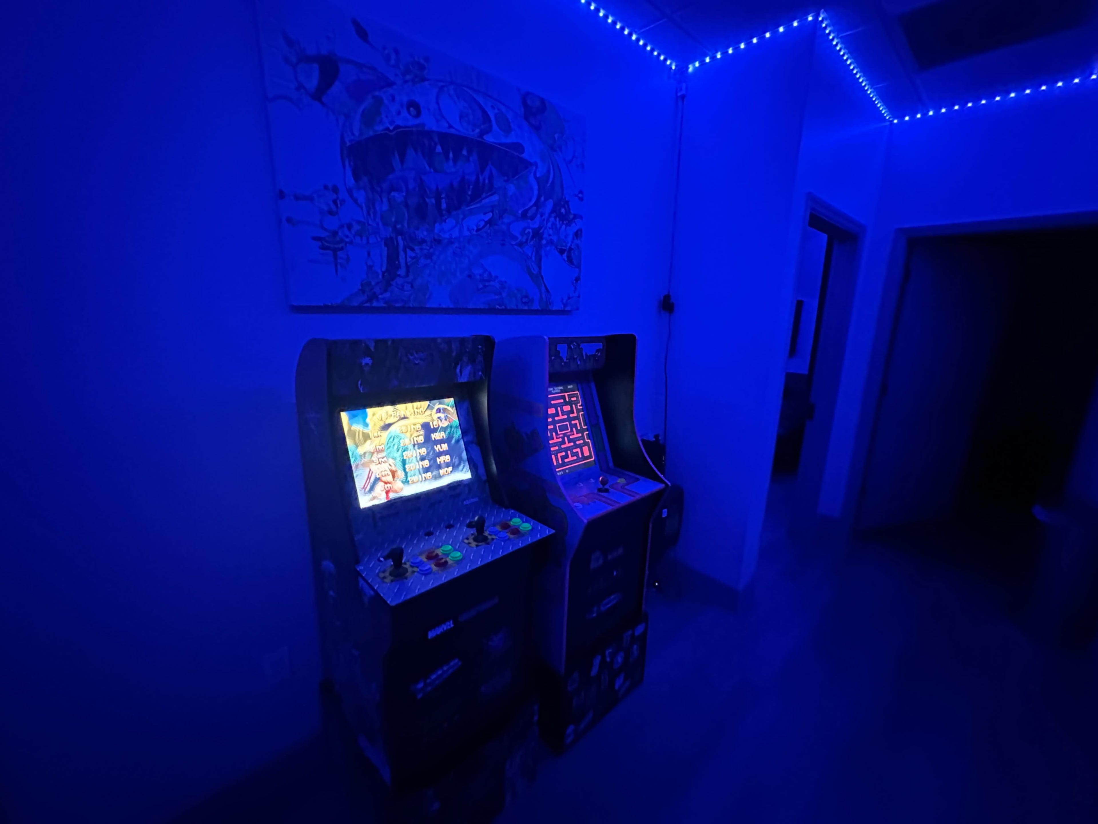 Two illuminated arcade game machines against a blue-lit wall, with a colorful artwork hanging above them.