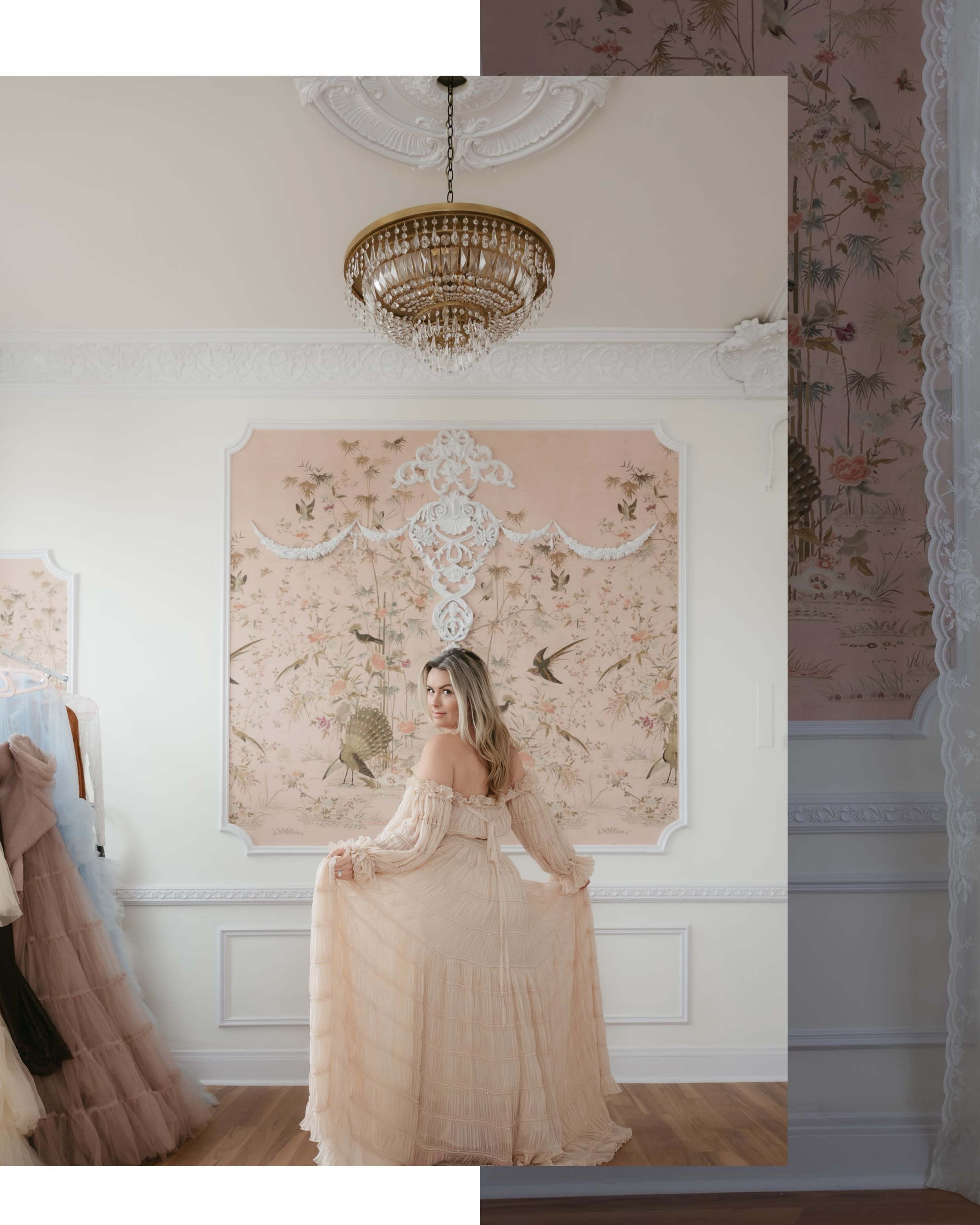 A woman in a flowing, off-the-shoulder dress poses in a elegantly decorated room with floral wallpaper and a chandelier.