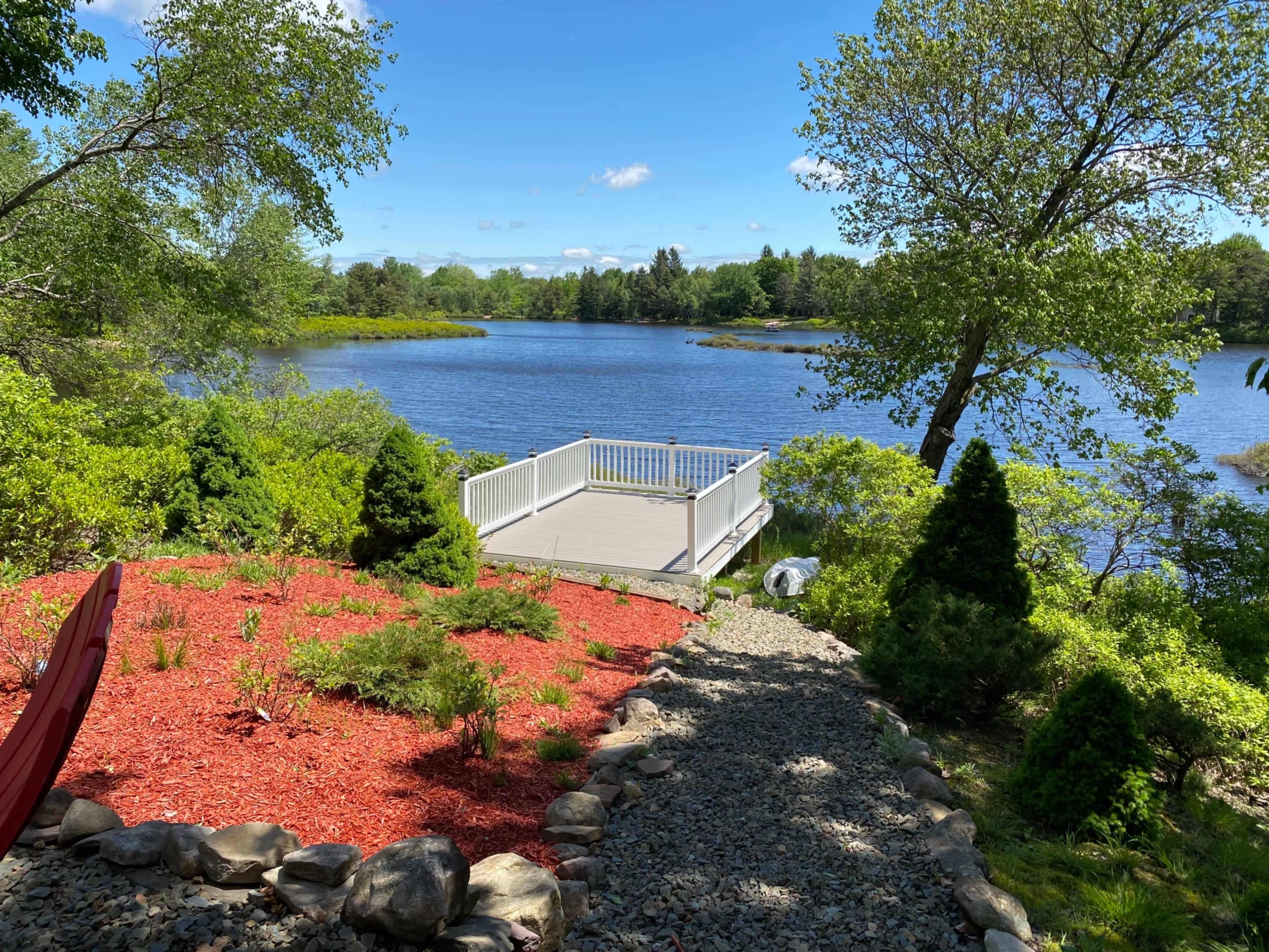The scene features a landscaped area with a gravel path leading to a wooden deck overlooking a calm lake surrounded by trees and greenery.