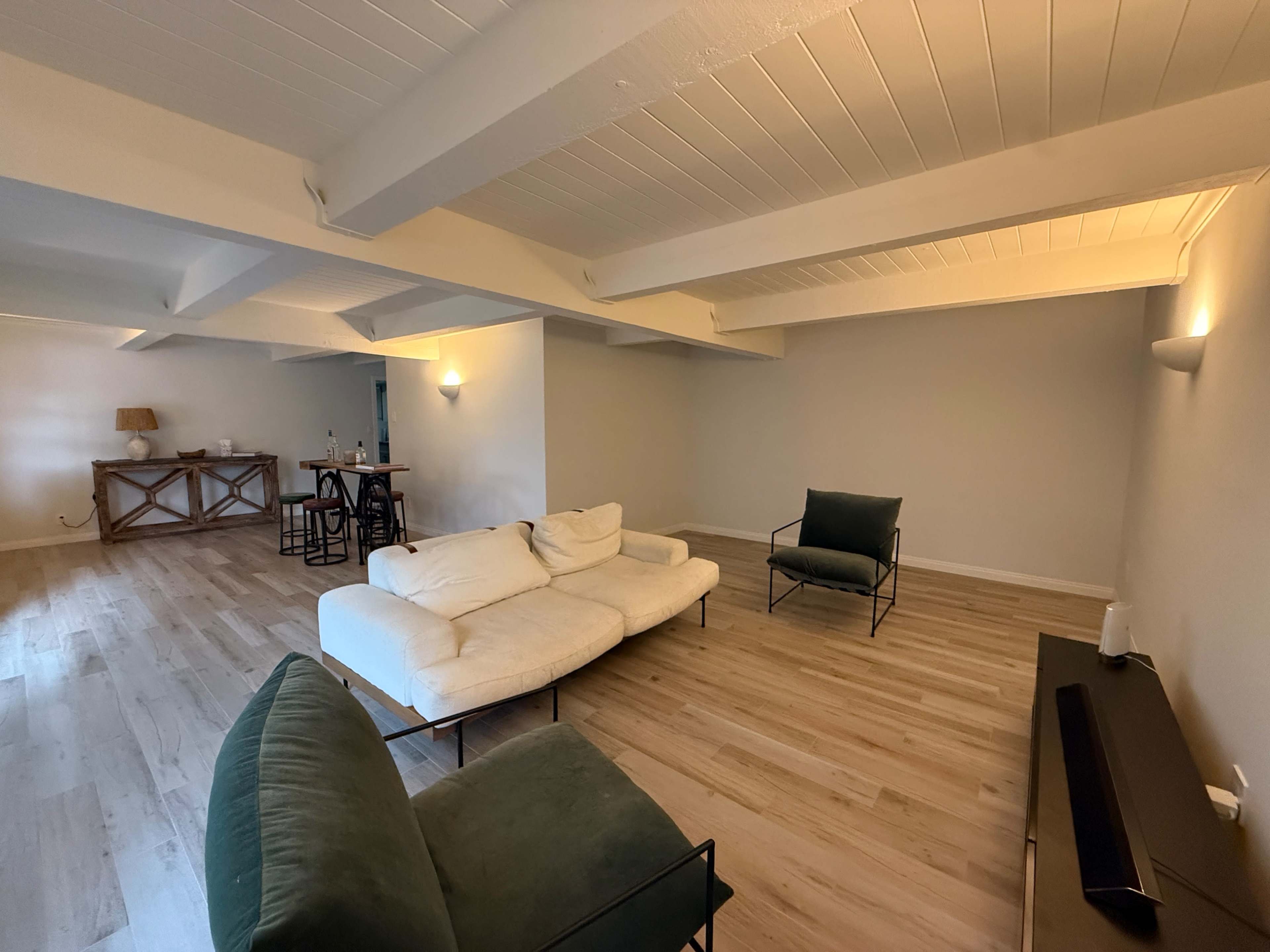 The image shows a spacious living area with a light-colored sofa, two green chairs, and wooden flooring, illuminated by soft lighting.