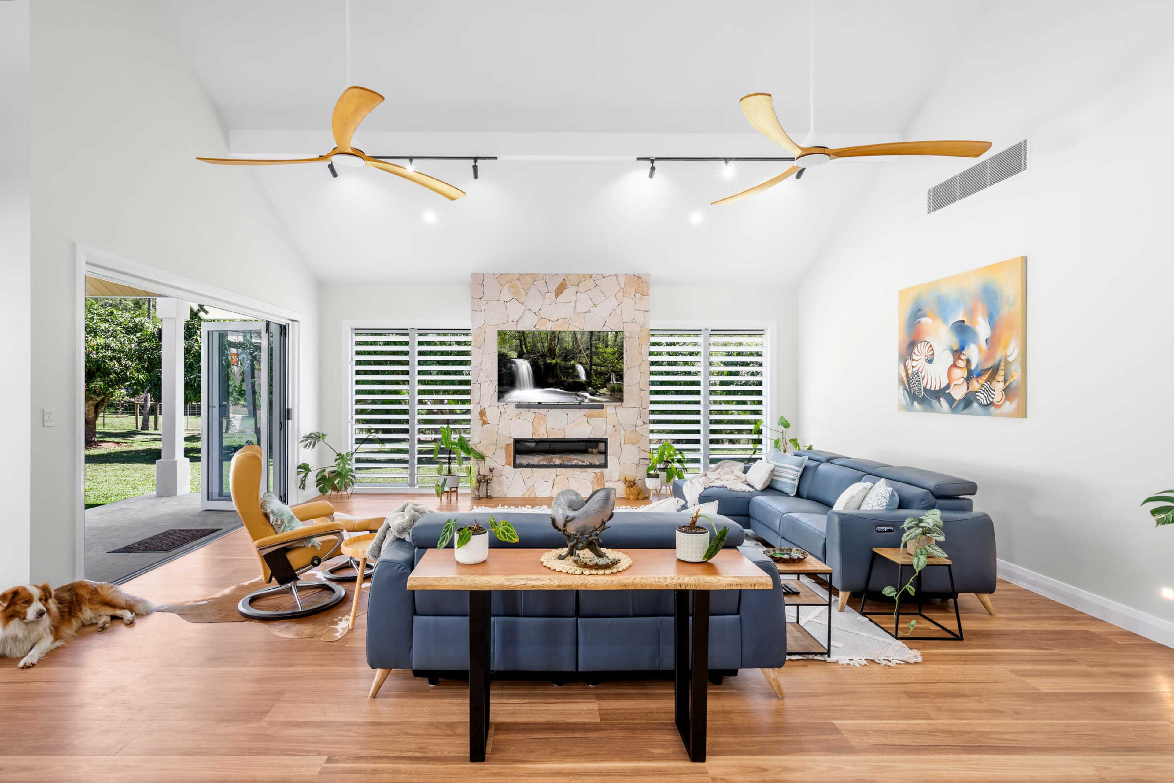 A spacious living room features a blue sectional sofa, a wooden coffee table with decorative items, and a stone fireplace beneath a wall-mounted television, with large windows that provide a view of the outdoor greenery.