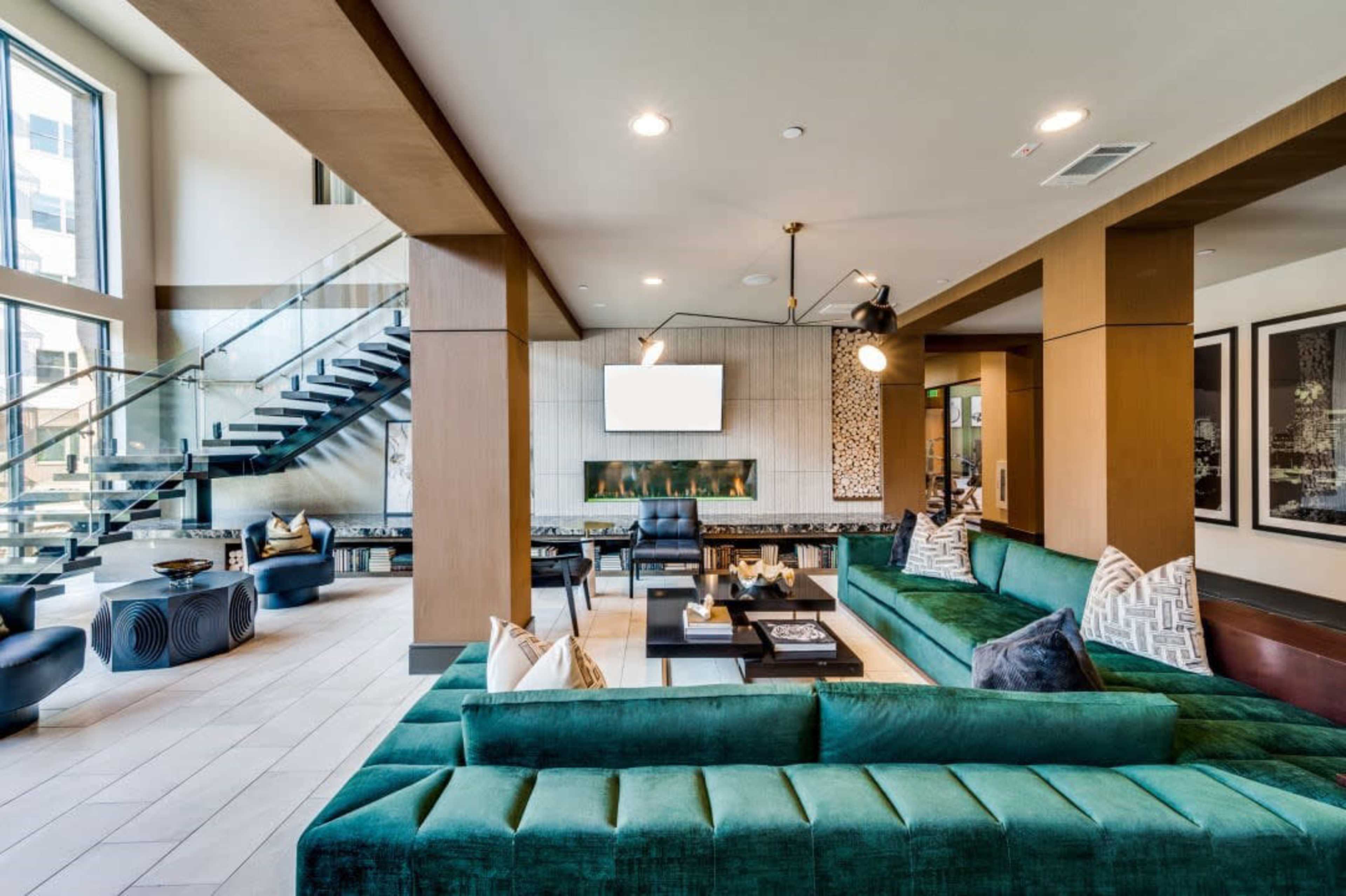 A modern interior lounge area featuring a green sectional sofa, a fireplace, a television mounted on the wall, and a staircase leading to an upper level.