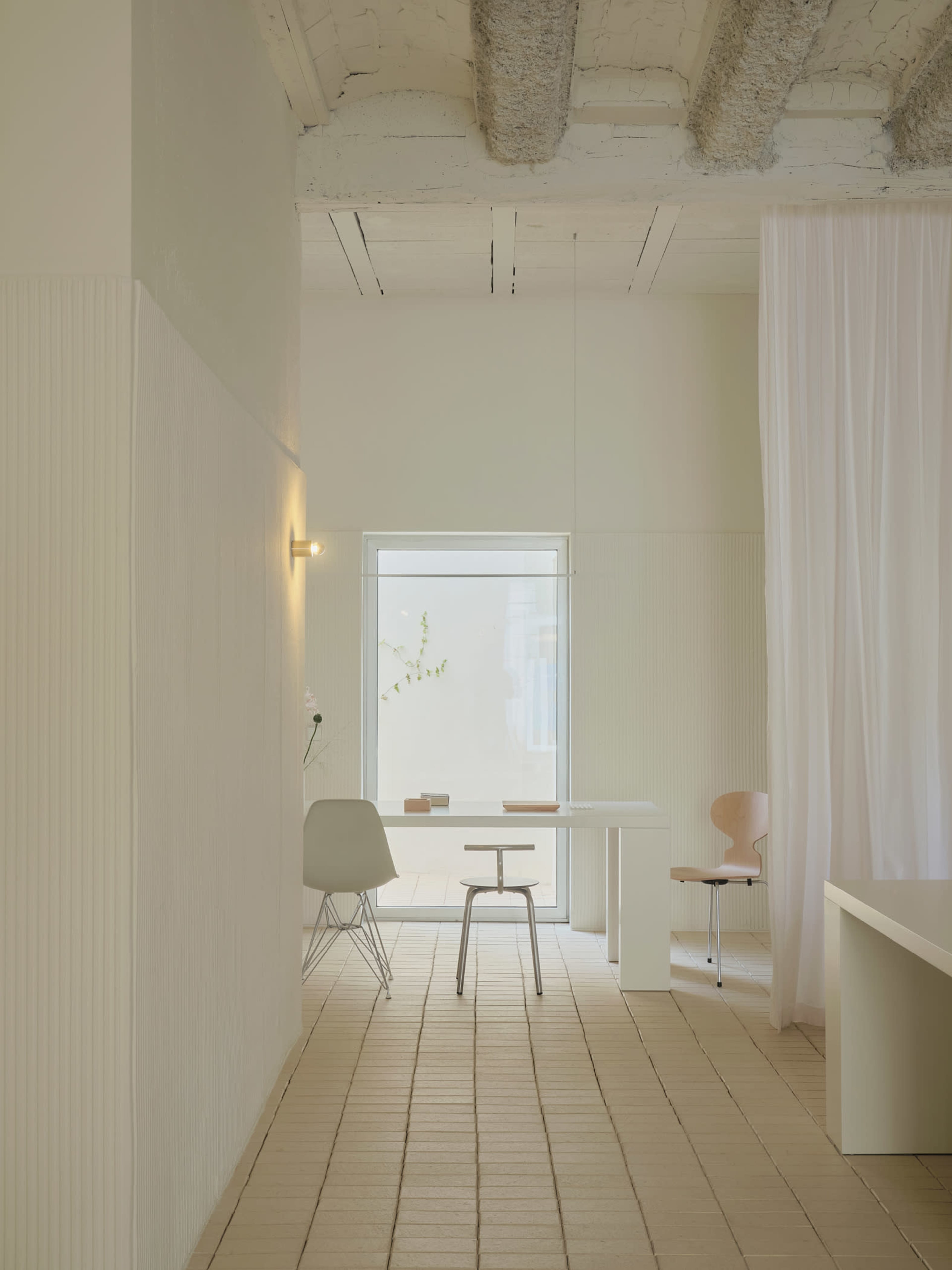The image shows a minimalist interior space with light walls, a wooden floor, and simple furniture including chairs and a table.