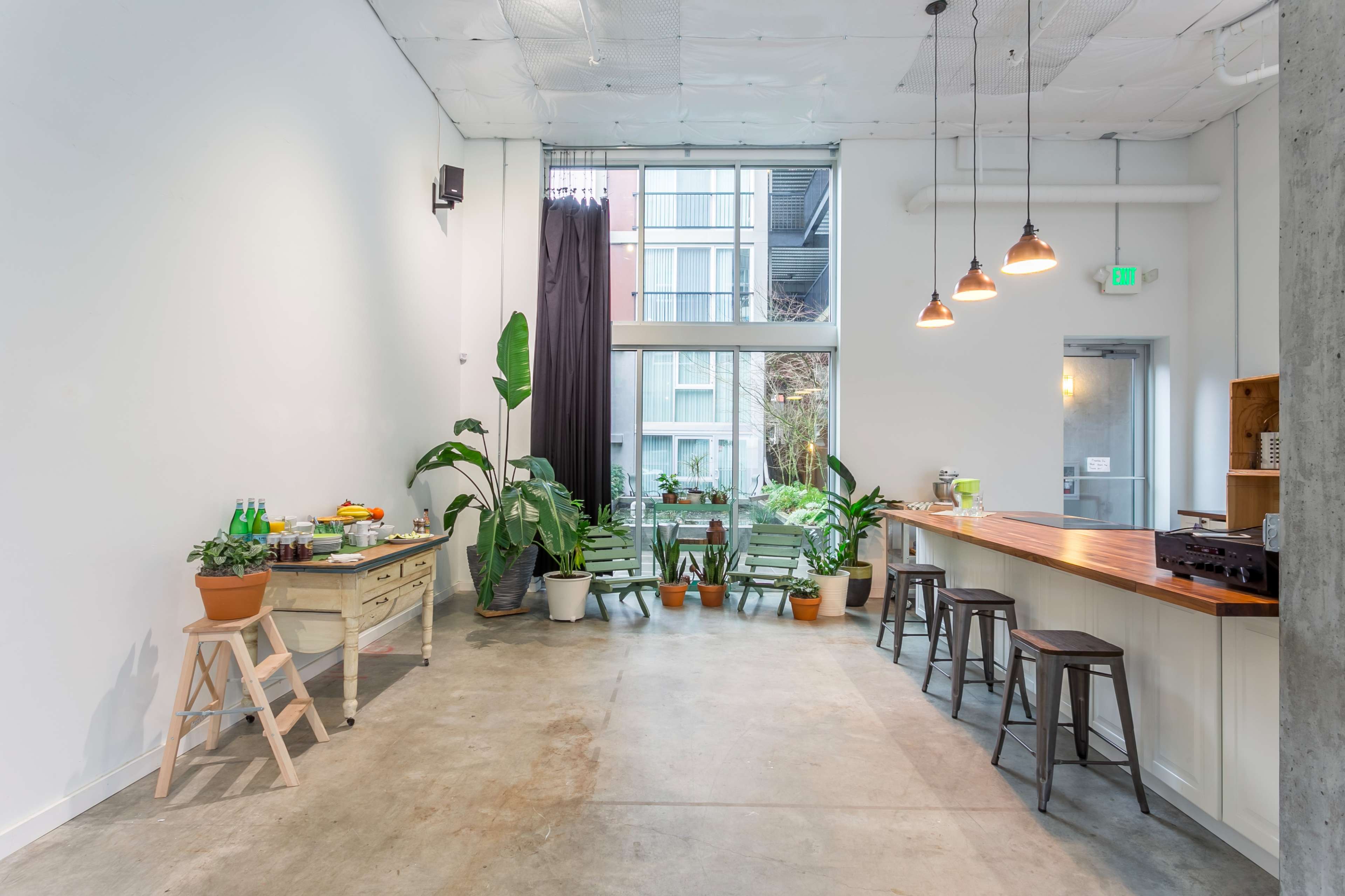 A minimalist interior space with a wooden counter, stools, plants in pots, and a large window letting in natural light.