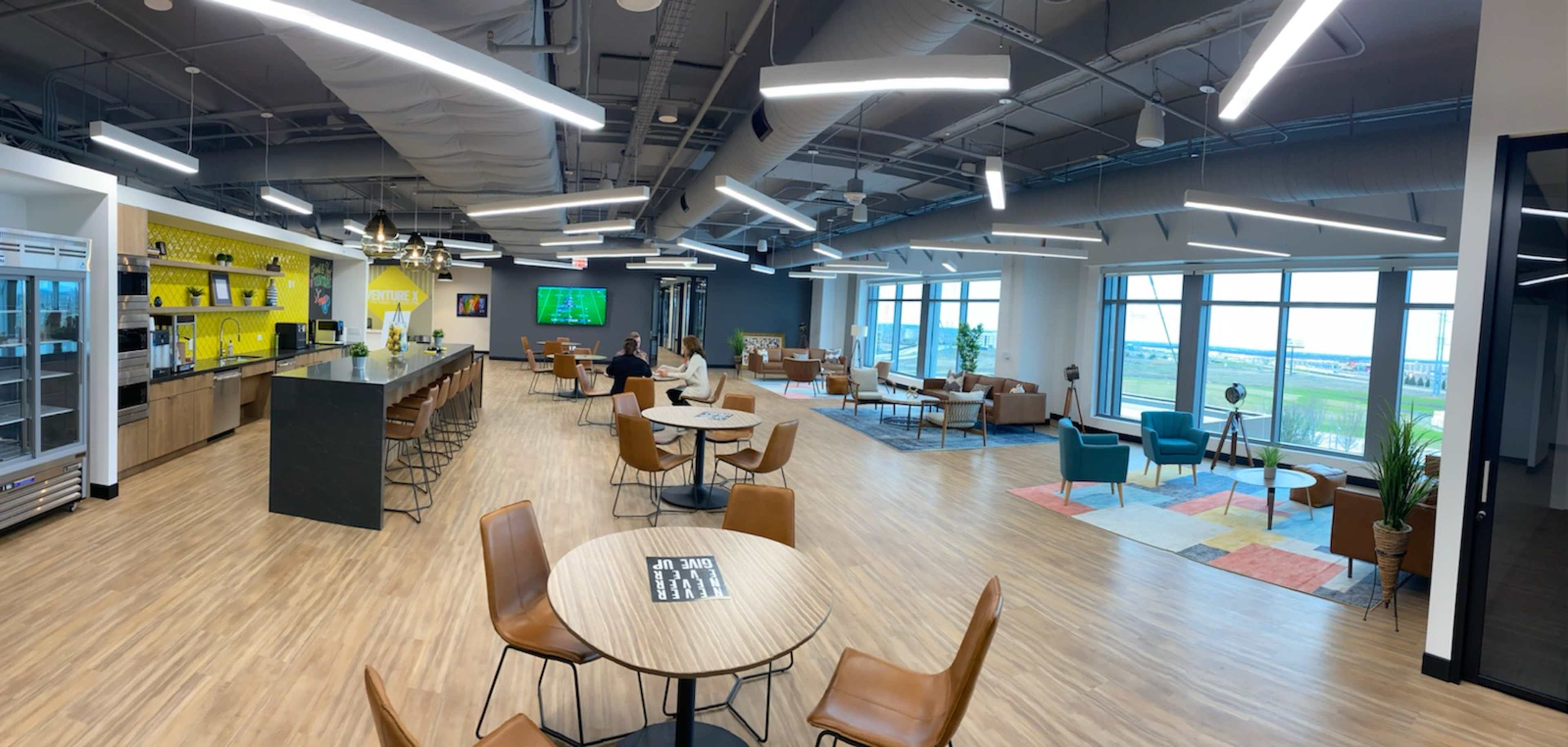 A modern office space with a kitchenette, seating areas, and large windows allowing natural light to enter.