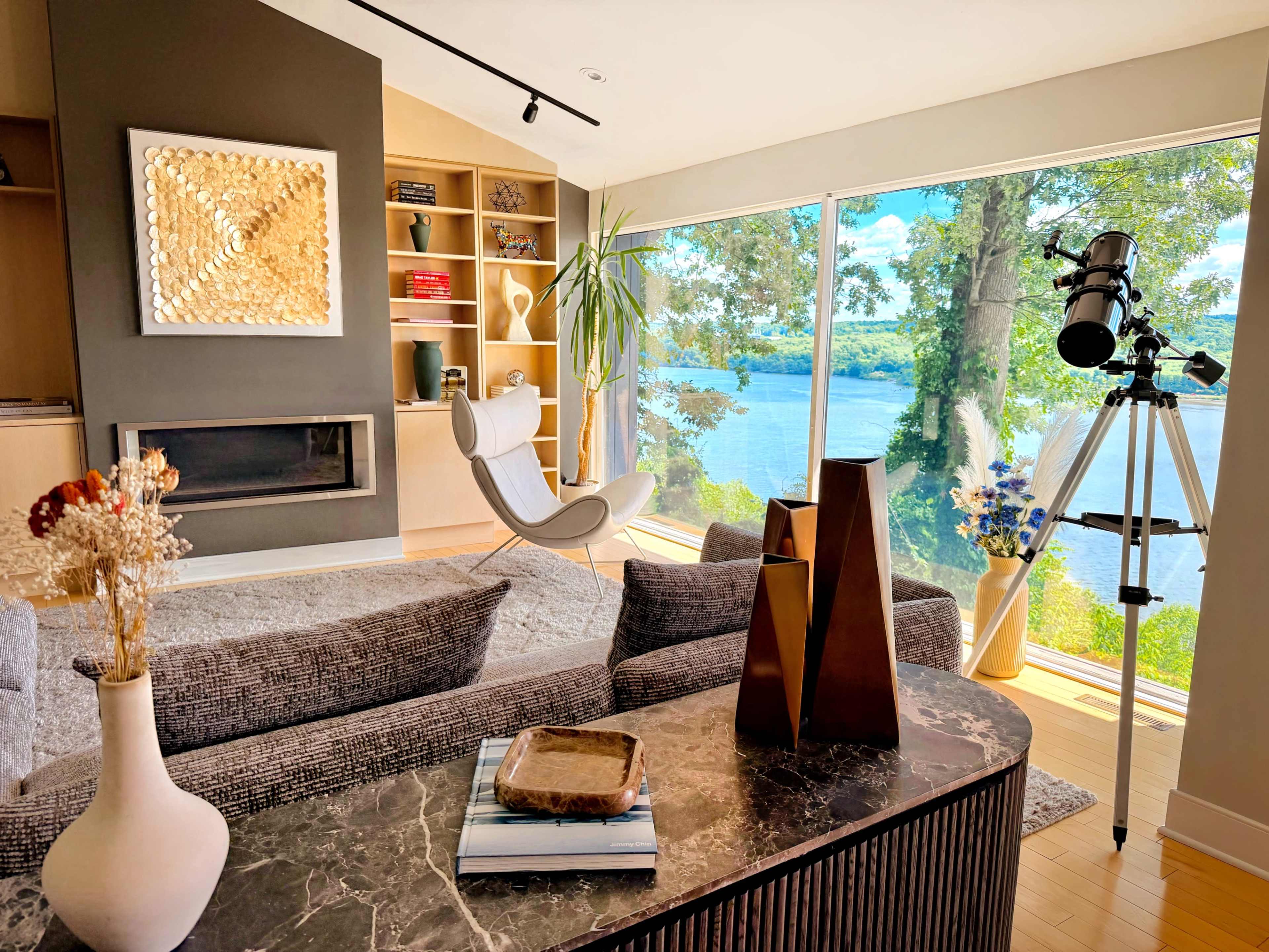 The image shows a modern living room with a large window overlooking a lake, featuring a telescope, a textured rug, and decorative items on a coffee table.