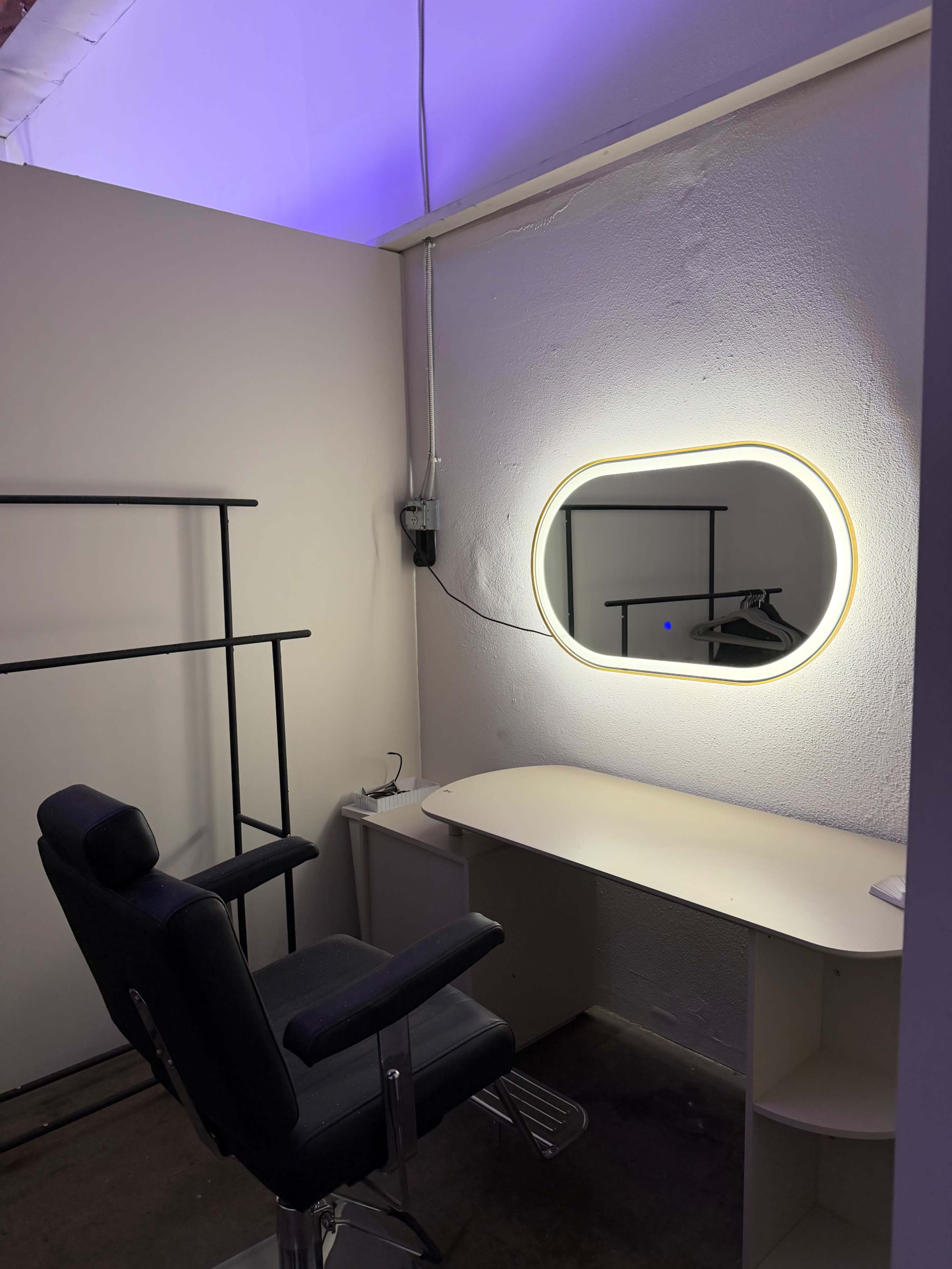 The image shows a minimalist room featuring a desk with an oval mirror and a black chair, illuminated by soft lighting from above.