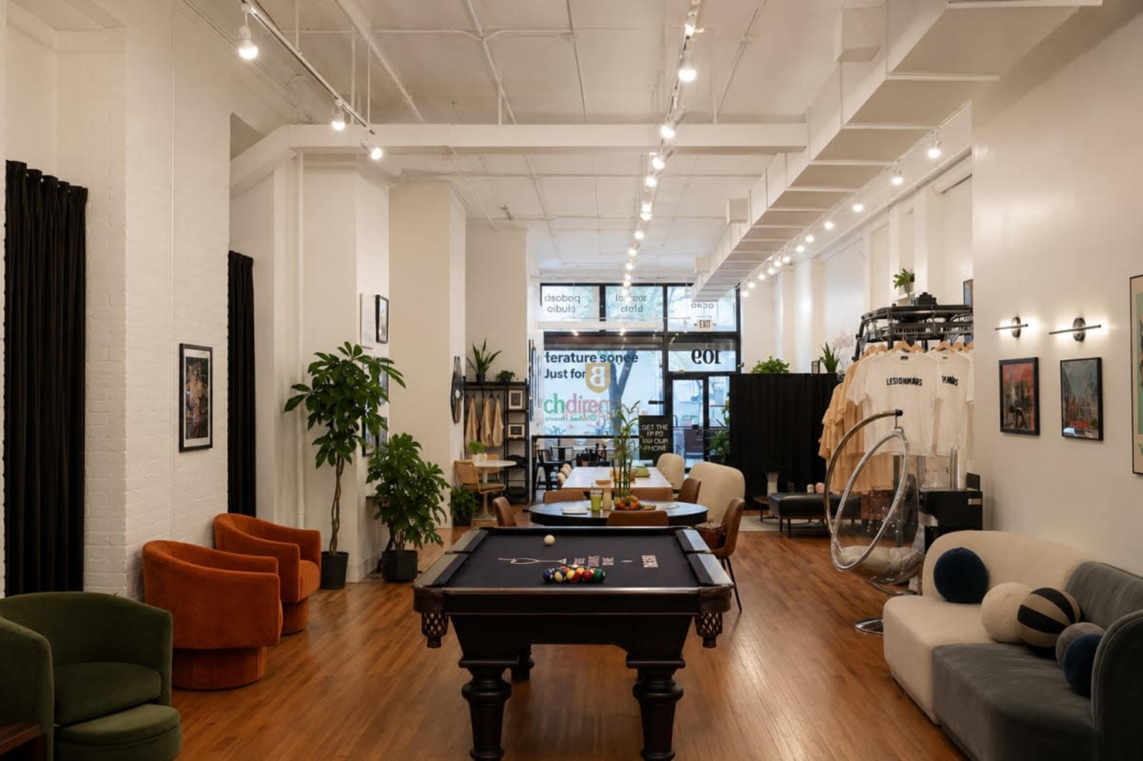 The image shows a spacious indoor area with a pool table, various seating arrangements, and display racks with clothing, illuminated by hanging lights.