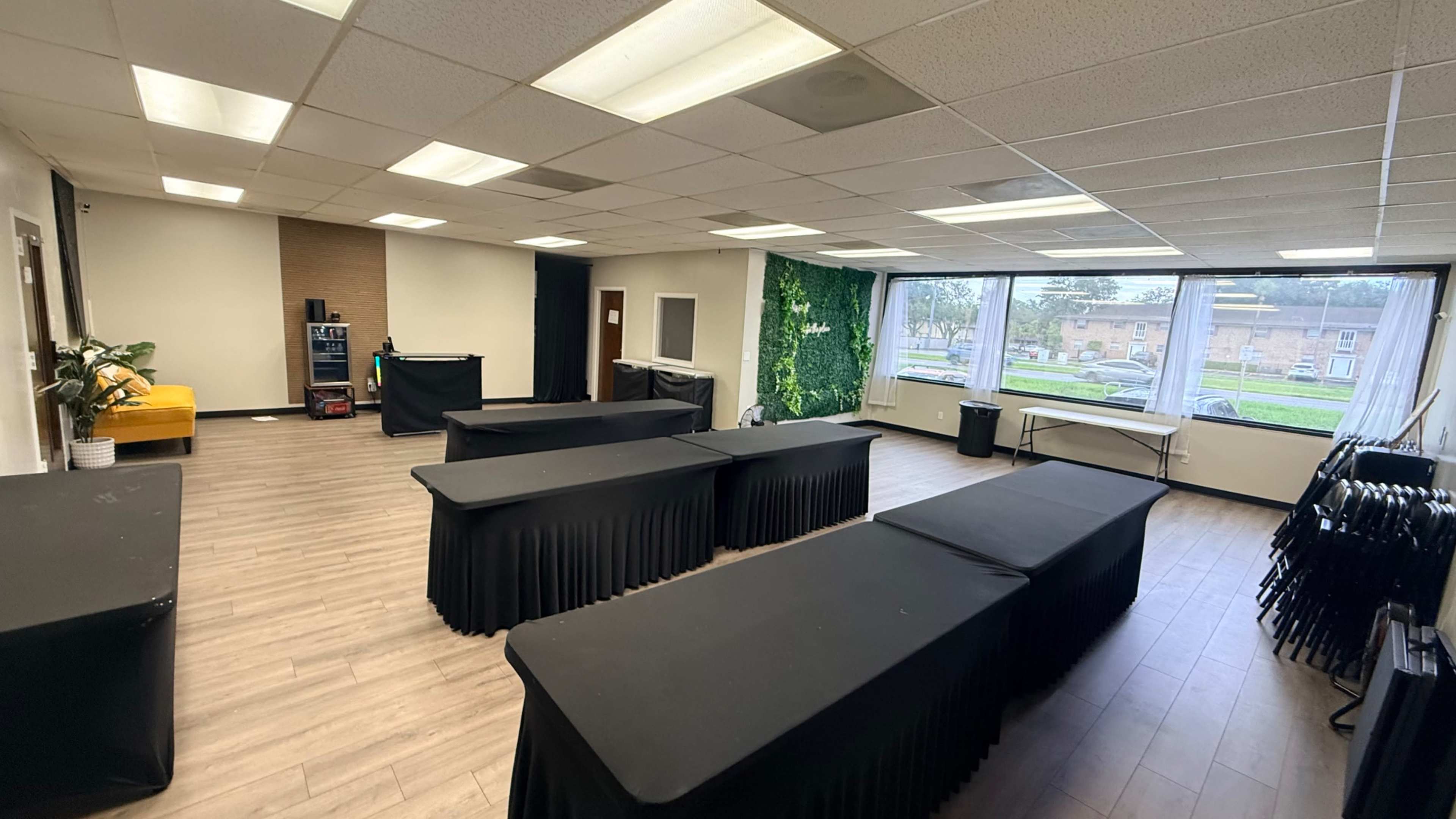 The image shows a spacious room with several black tables arranged in the center, a green wall feature, and large windows letting in natural light.