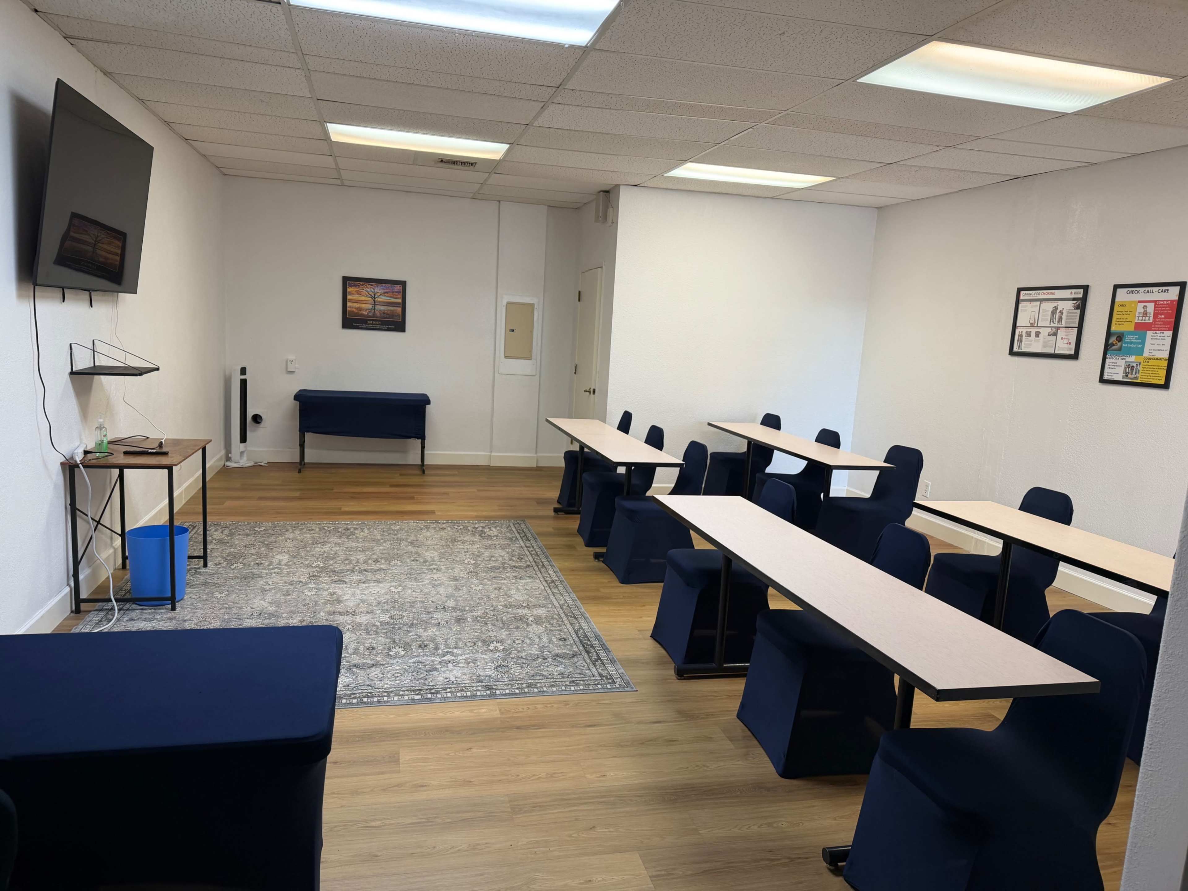 The image shows a bright, empty meeting room featuring several tables and chairs arranged in rows, along with a television mounted on the wall.
