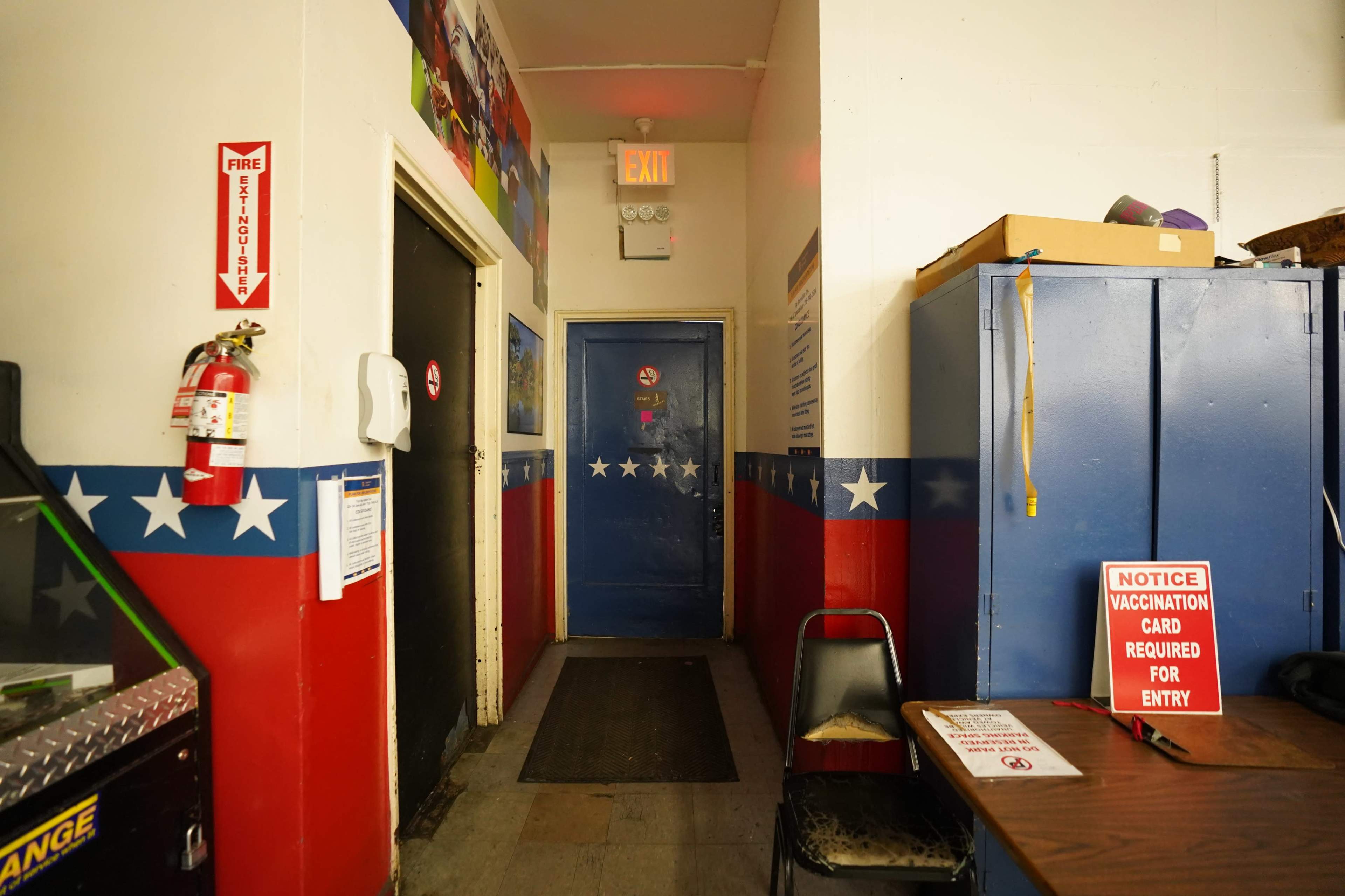 The image shows a corridor with red, white, and blue walls, a fire extinguisher mounted near the entrance, and signs indicating entry requirements and safety notices.