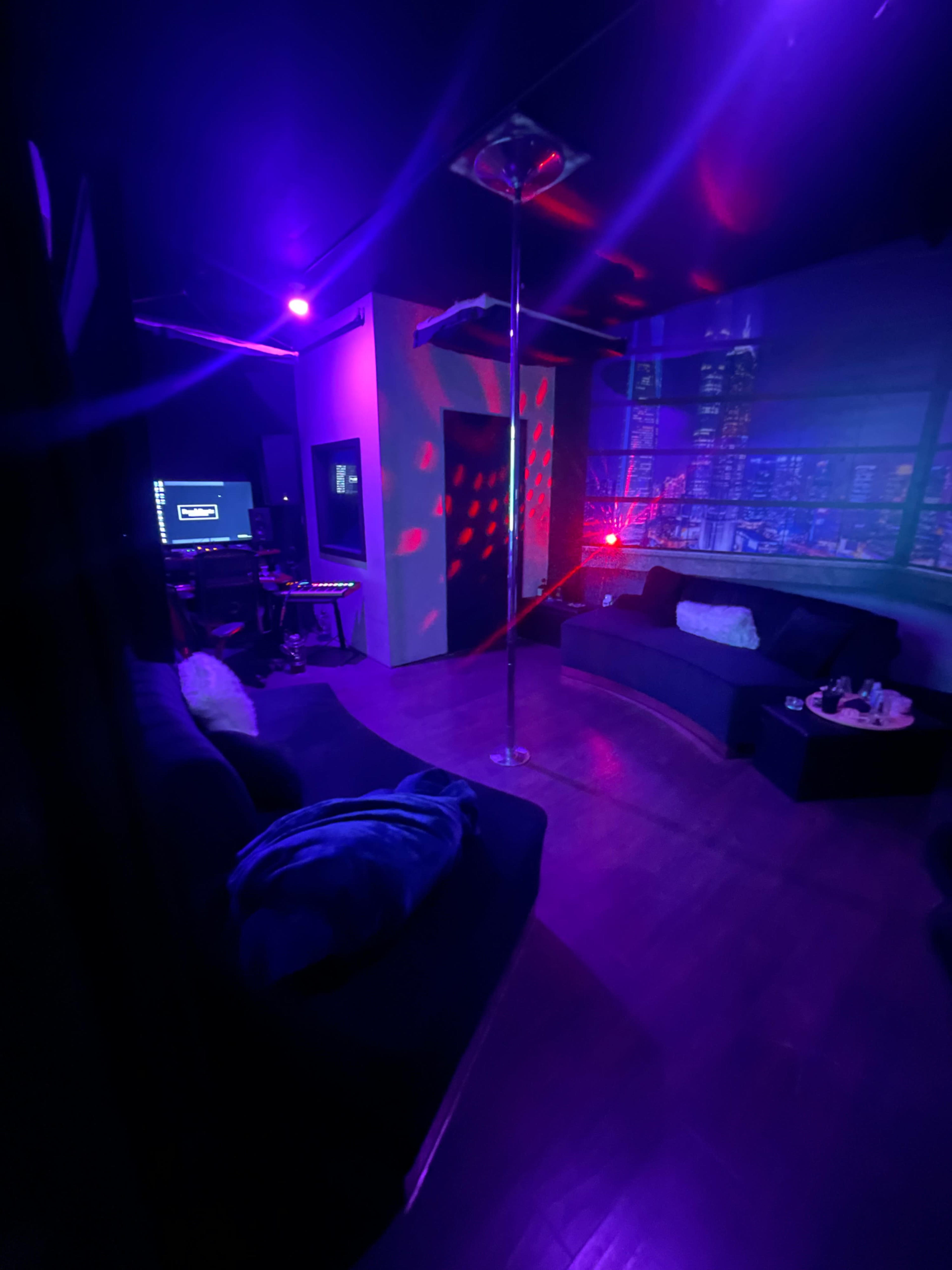 The image shows a dimly lit lounge with purple lighting, a stage featuring a pole, and seating areas, creating a nightclub atmosphere.