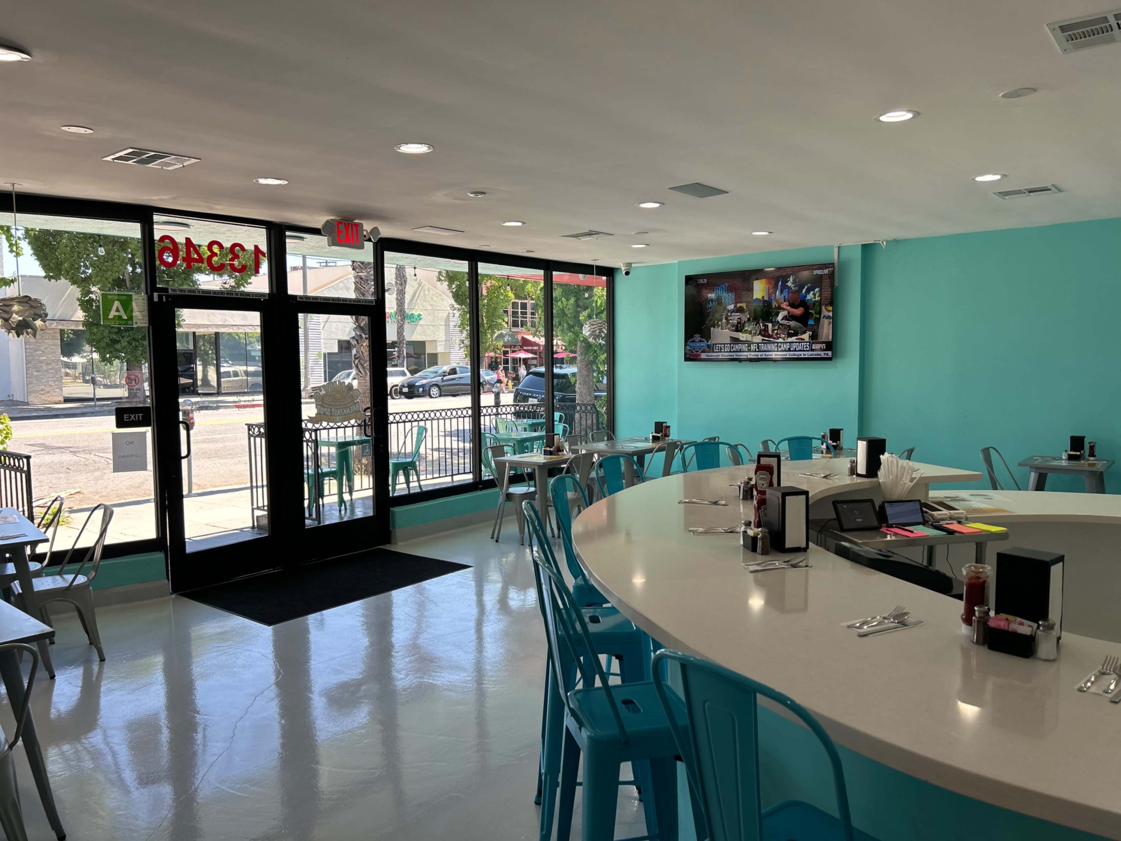The image shows a brightly colored cafe with turquoise walls, a large round table in the center, and large windows providing a view of the street outside.