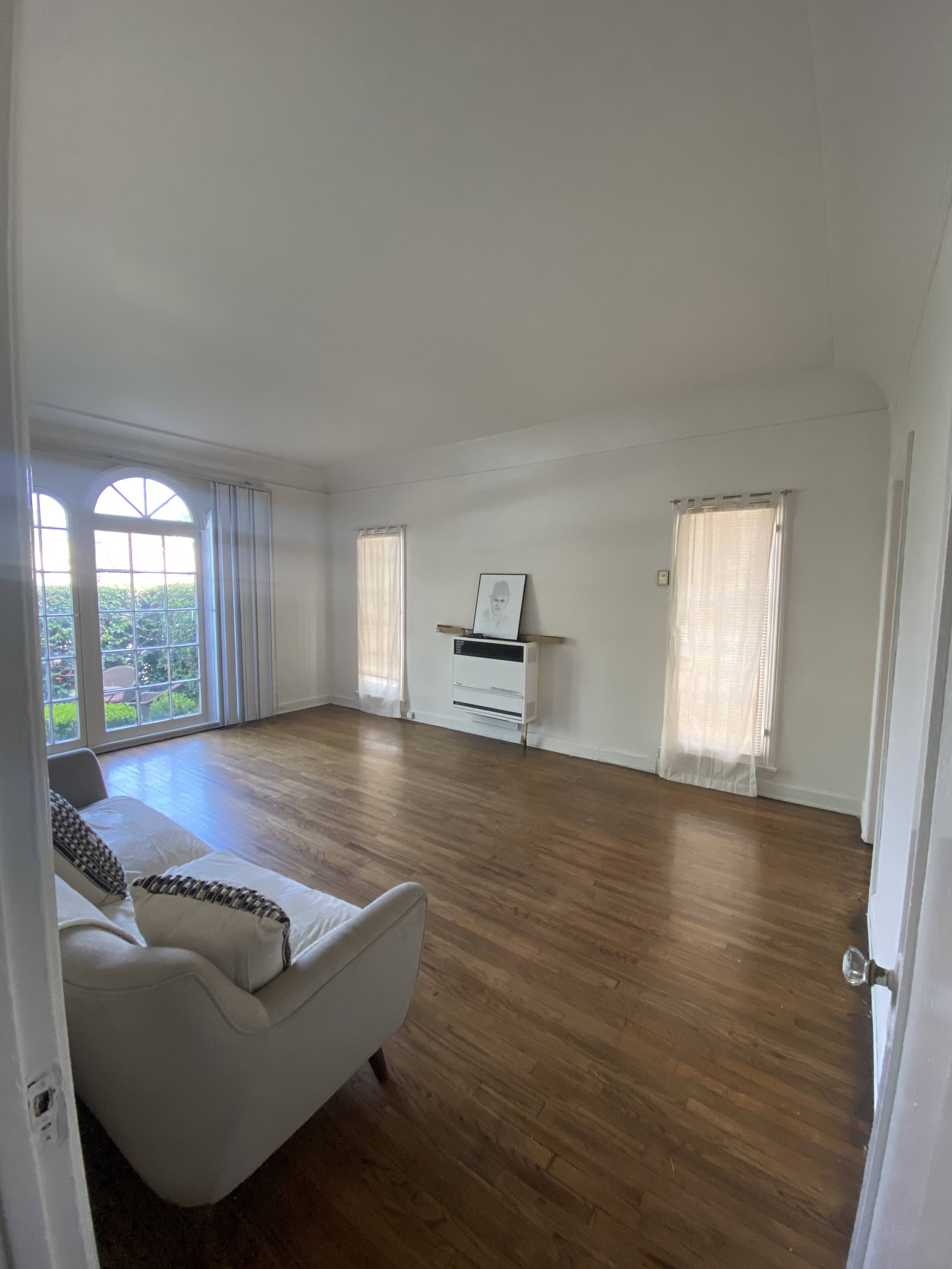 The image shows a spacious, minimally furnished living room with a hardwood floor and large windows allowing natural light.