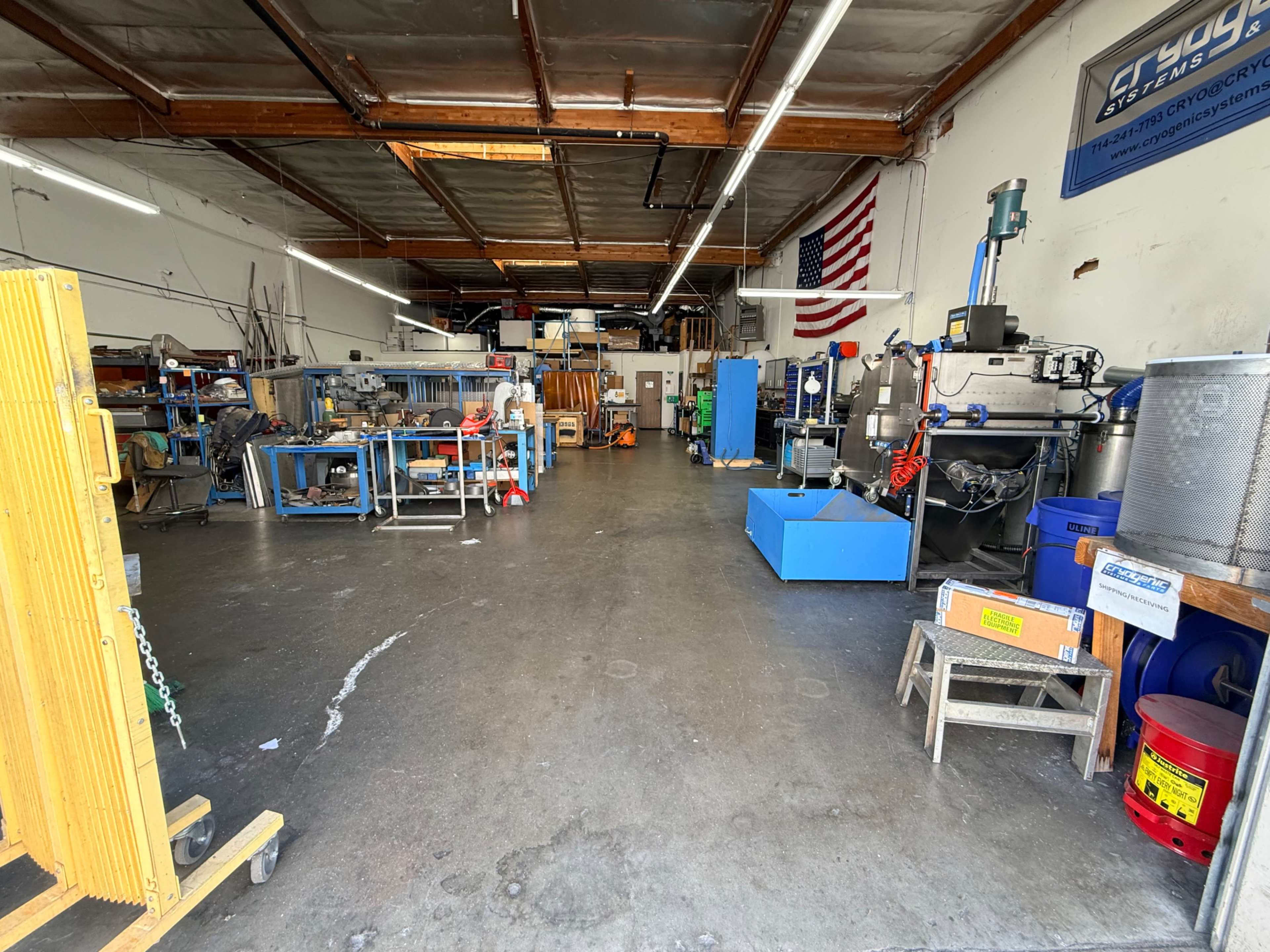 The image shows a spacious industrial workshop with various machinery and equipment arranged on the floor, along with an American flag displayed on the wall.