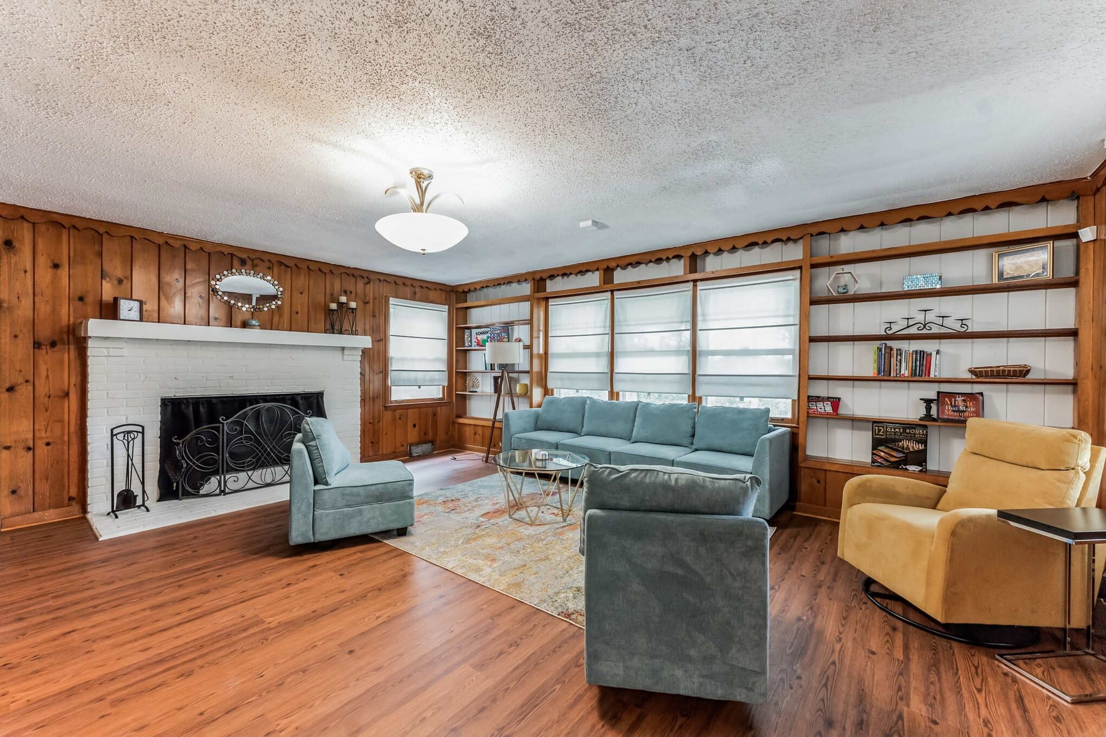 The living room features a white brick fireplace surrounded by wooden paneling, with a light blue sofa set, a glass coffee table, and built-in shelves filled with books and decorative items.