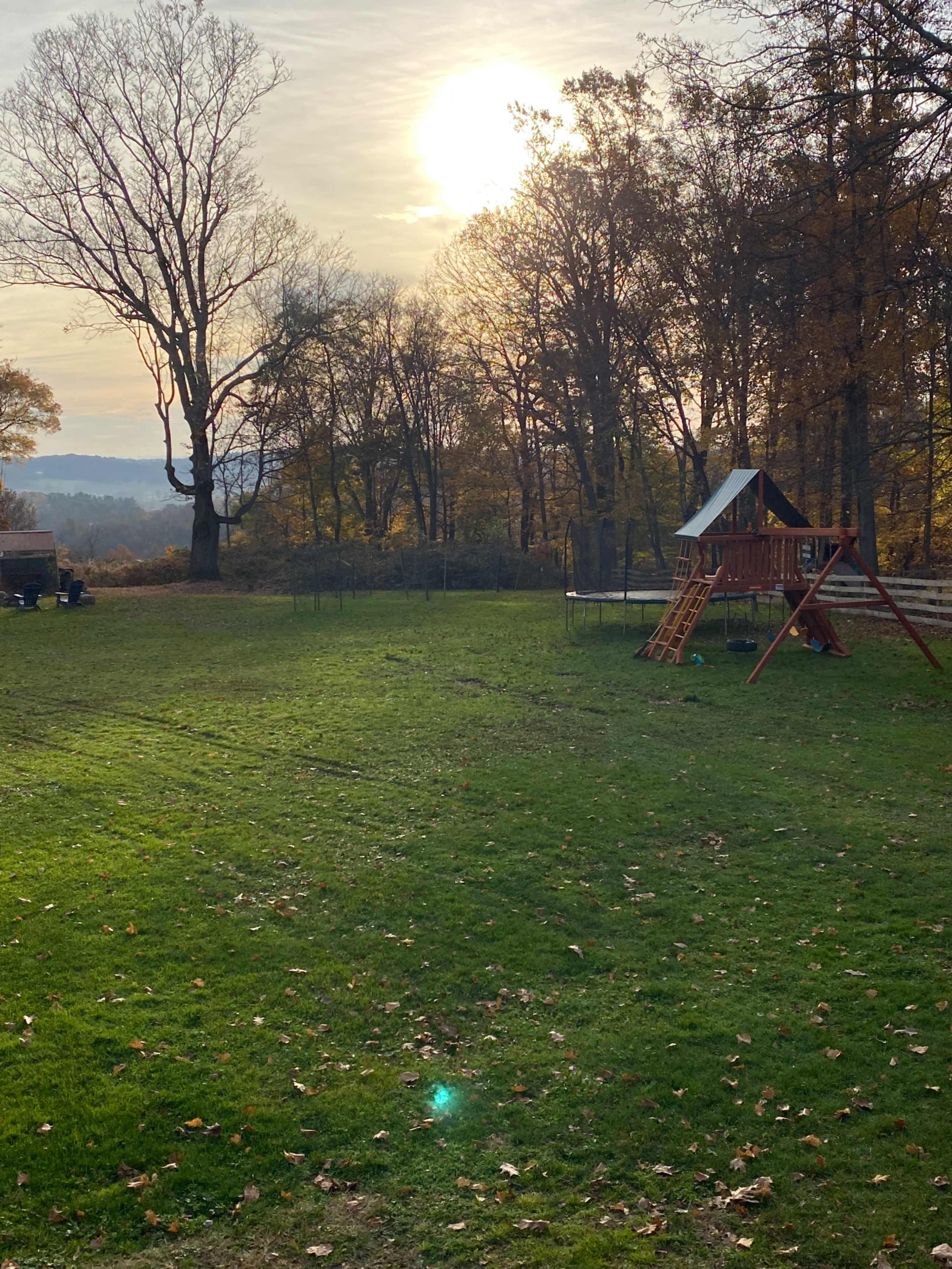 A playground set stands on a grassy lawn under a setting sun, surrounded by trees with autumn leaves.