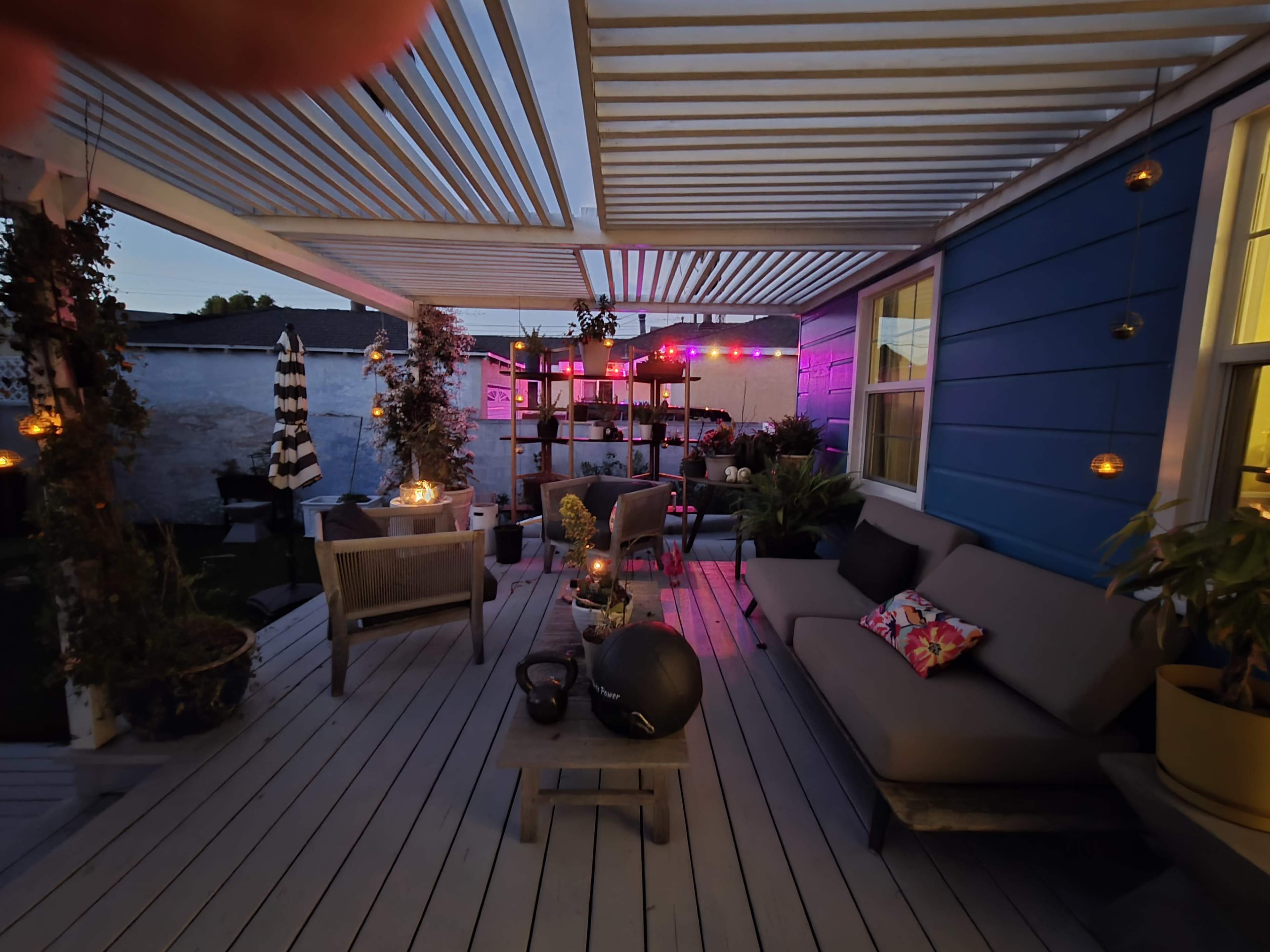 The image shows a cozy outdoor patio area with seating, a small table, plants, and colorful lights in the background.