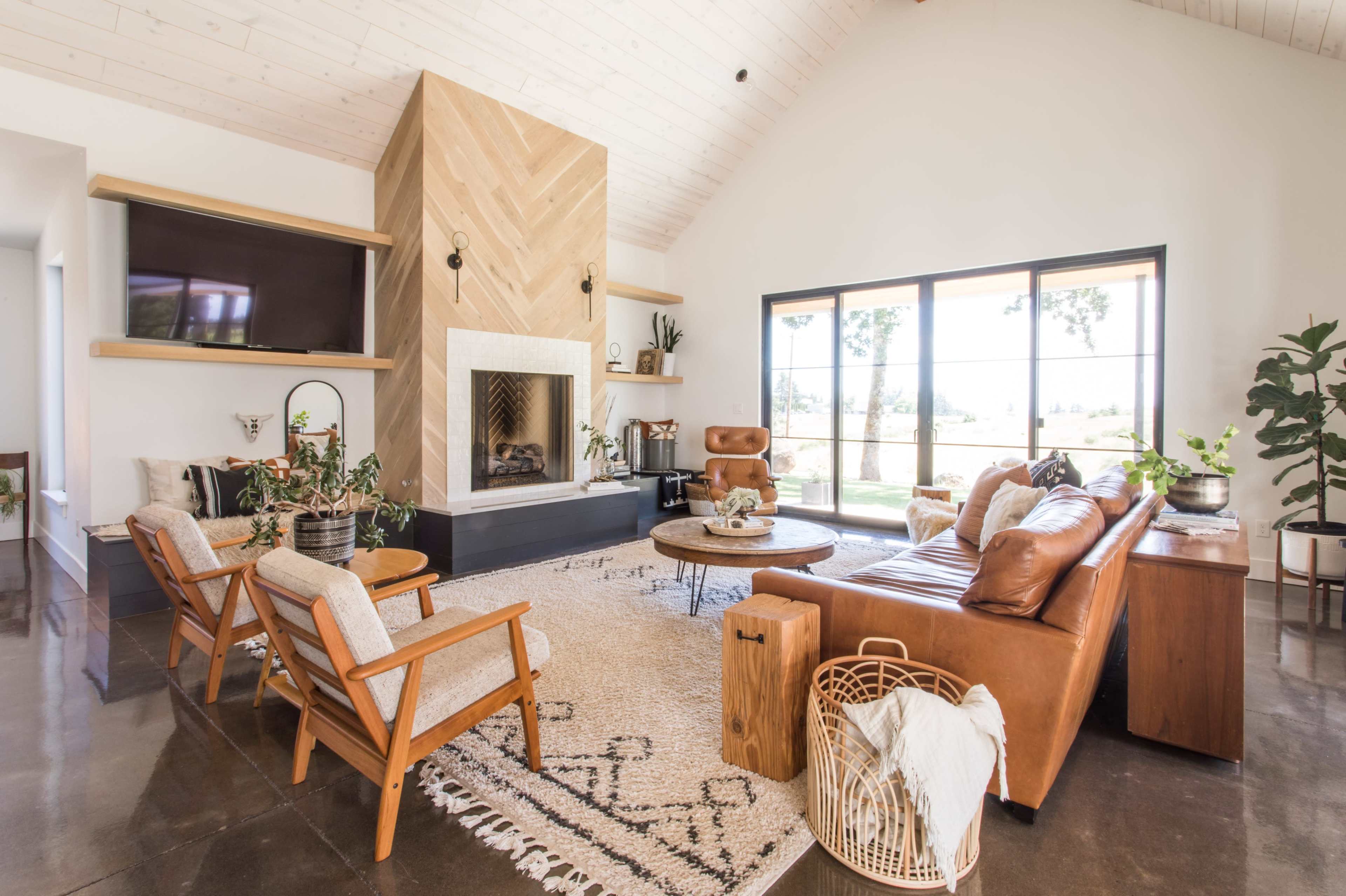 A modern living room with a large fireplace, a leather sofa, wooden chairs, and glass doors that lead to an outdoor view.