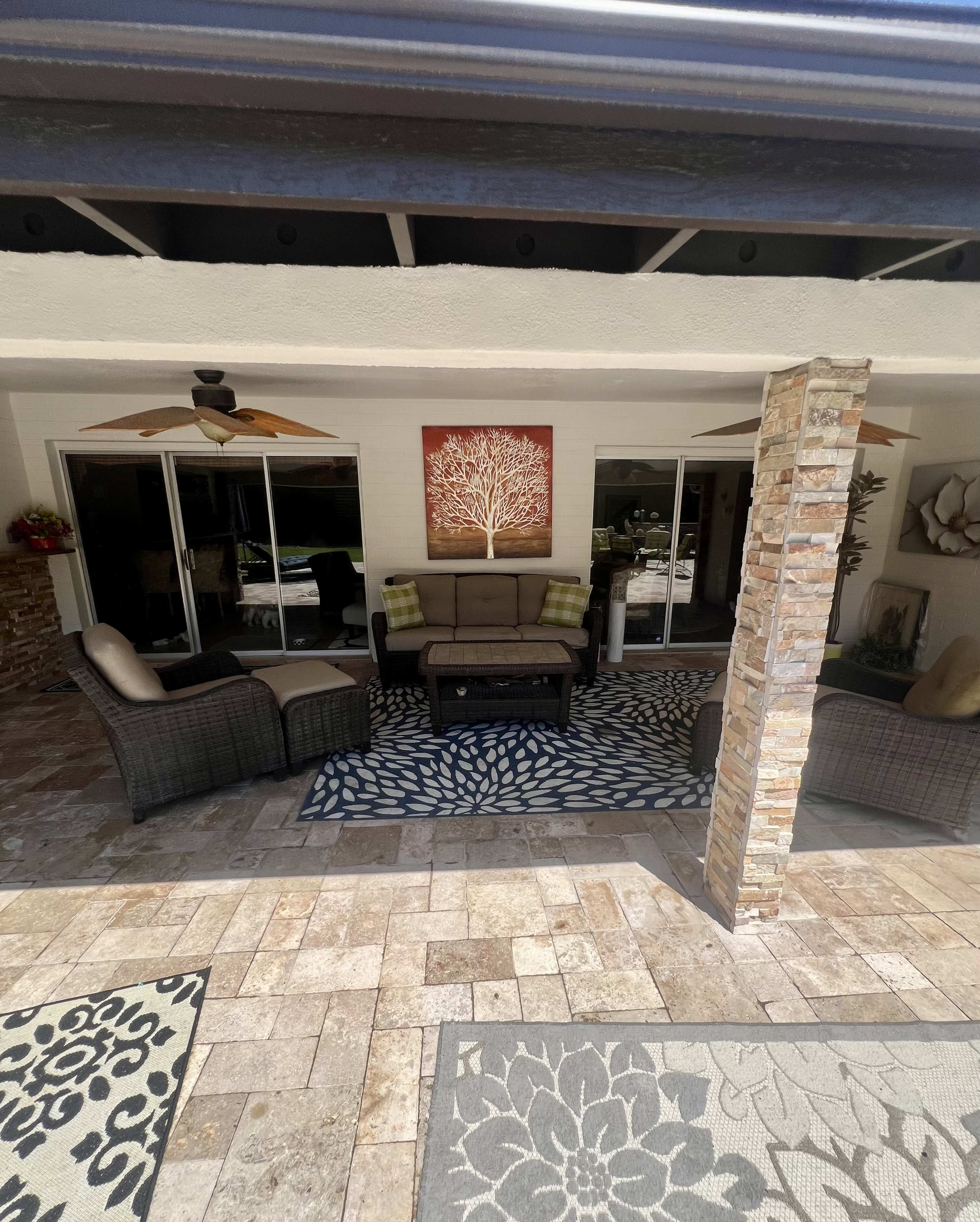 The image shows a covered outdoor patio with a seating arrangement of cushioned chairs and a coffee table, surrounded by decorative rugs and sliding glass doors.