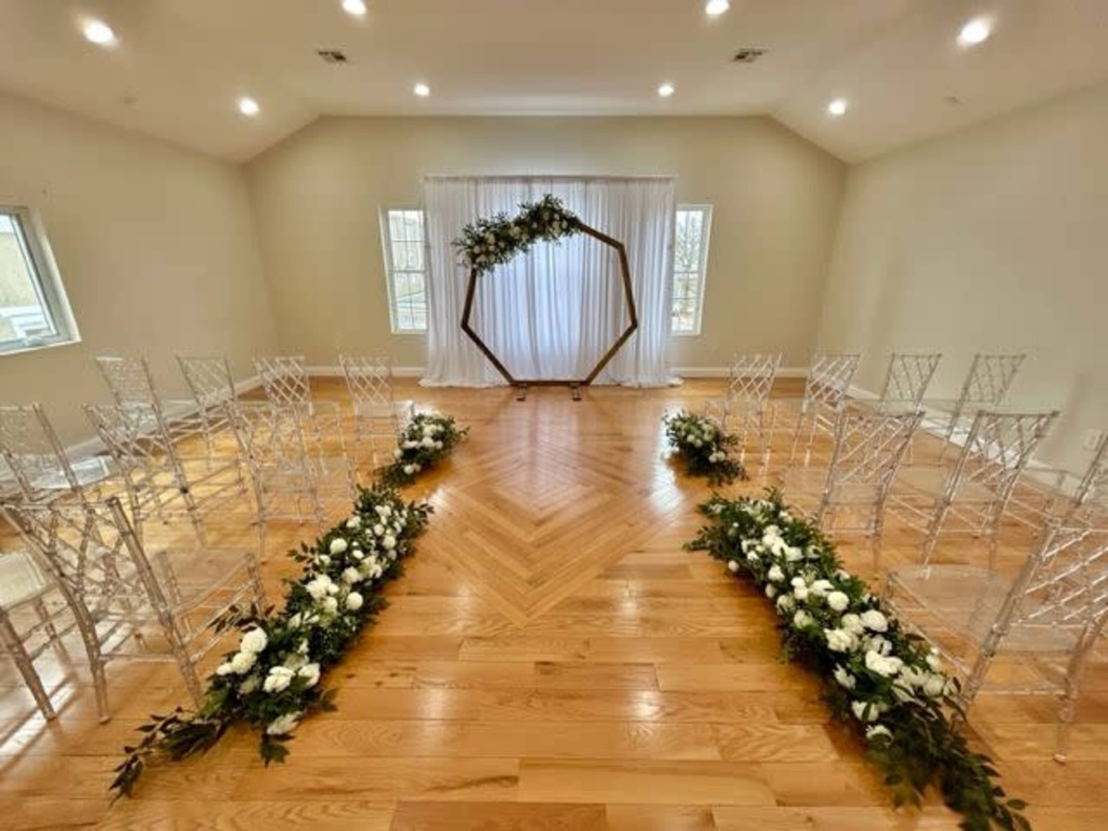 A ceremony space features an arch decorated with greenery and flowers, surrounded by rows of clear chairs and floral arrangements on the floor.