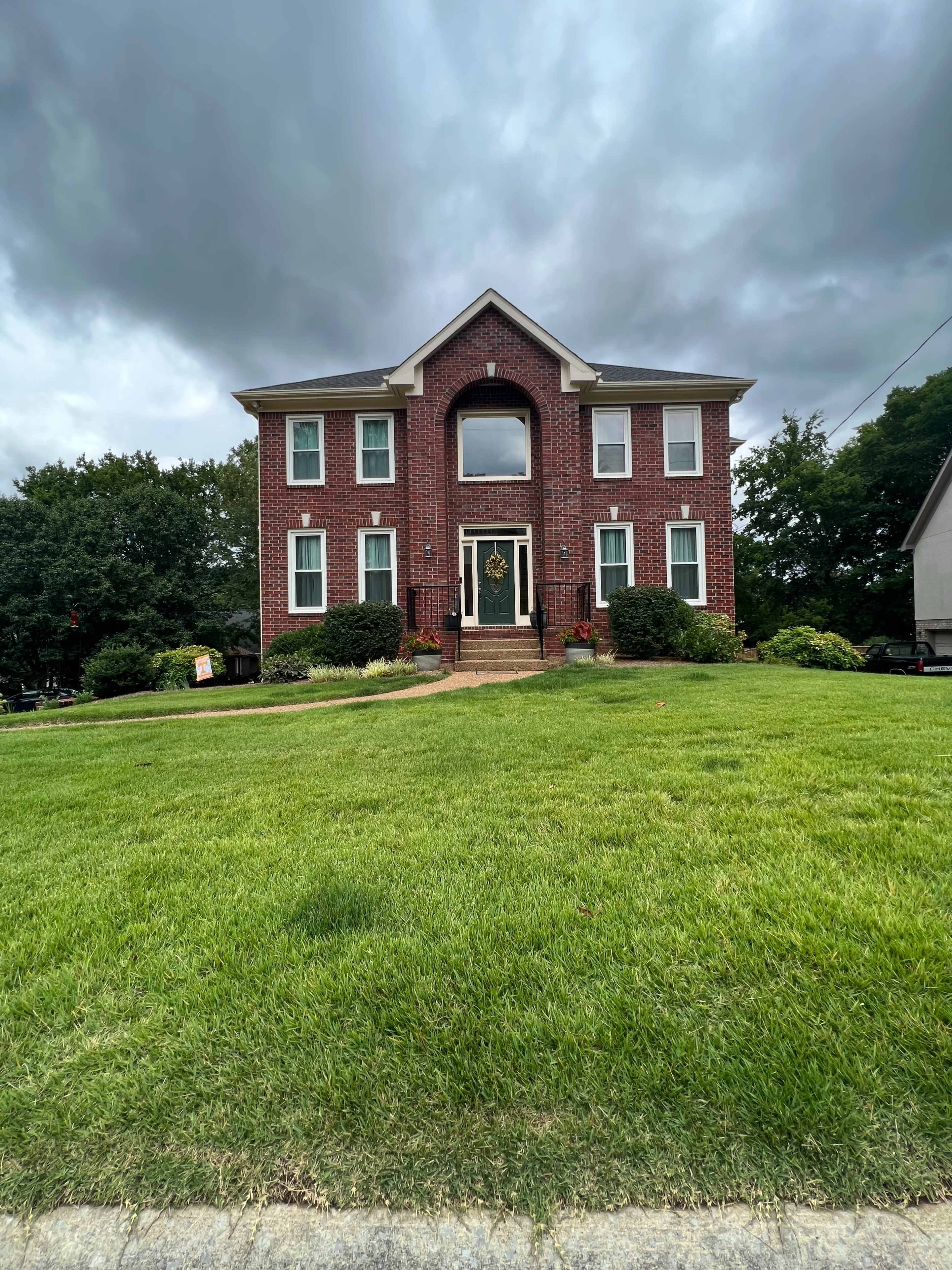A two-story brick house with a central entrance and a large front lawn is situated under a cloudy sky.