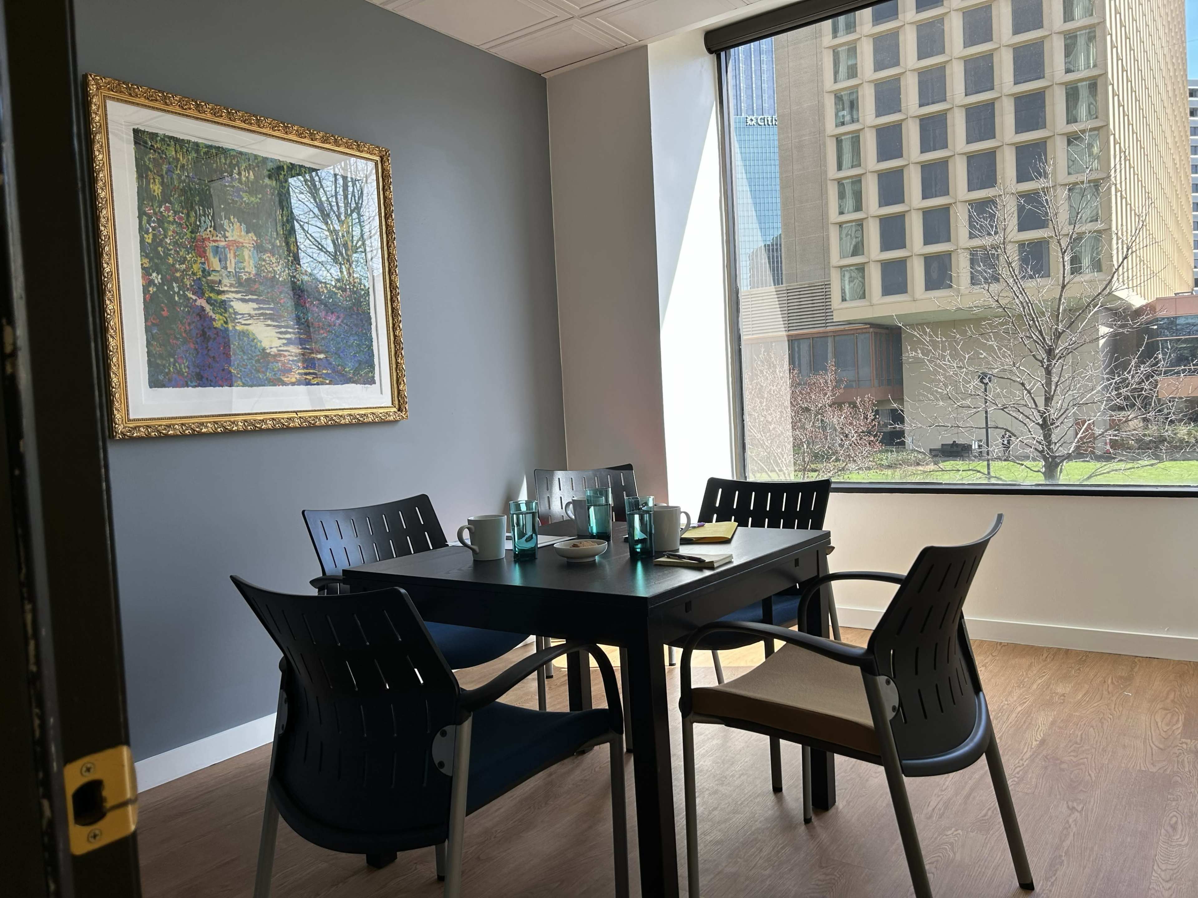 A small meeting room features a table set for a meal, surrounded by four chairs, with a large window providing a view of a building exterior and greenery outside.