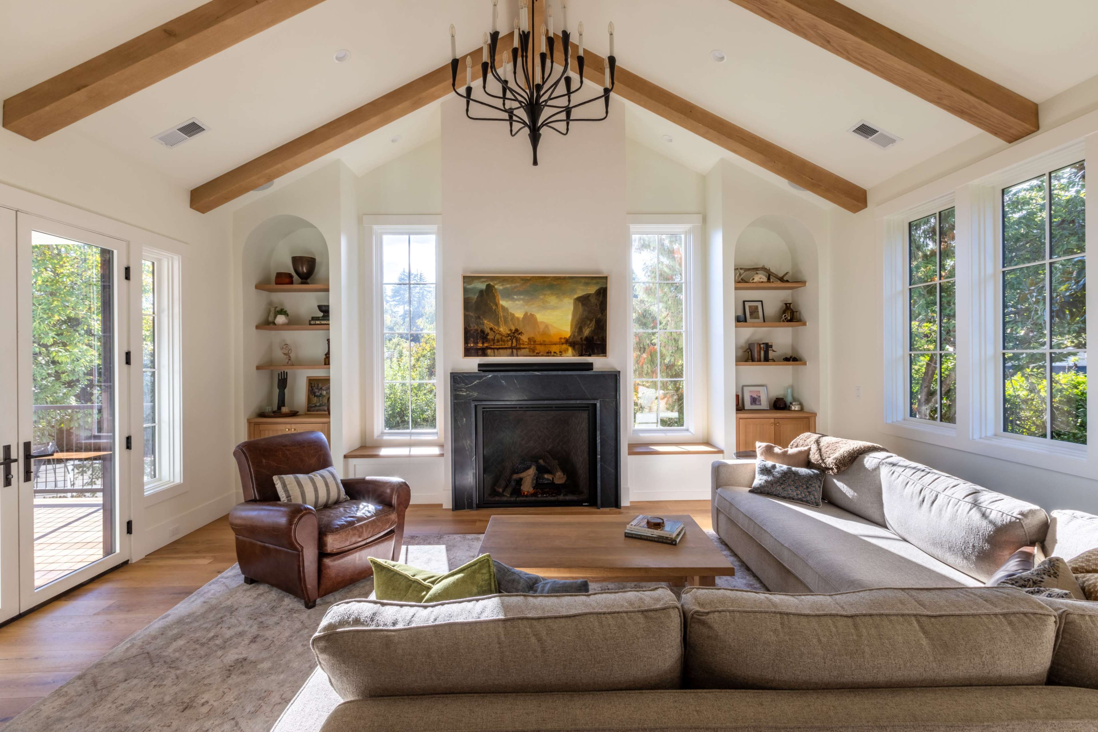 The image shows a bright living room with a high, wooden-beamed ceiling, a central fireplace, and large windows that overlook a green landscape.