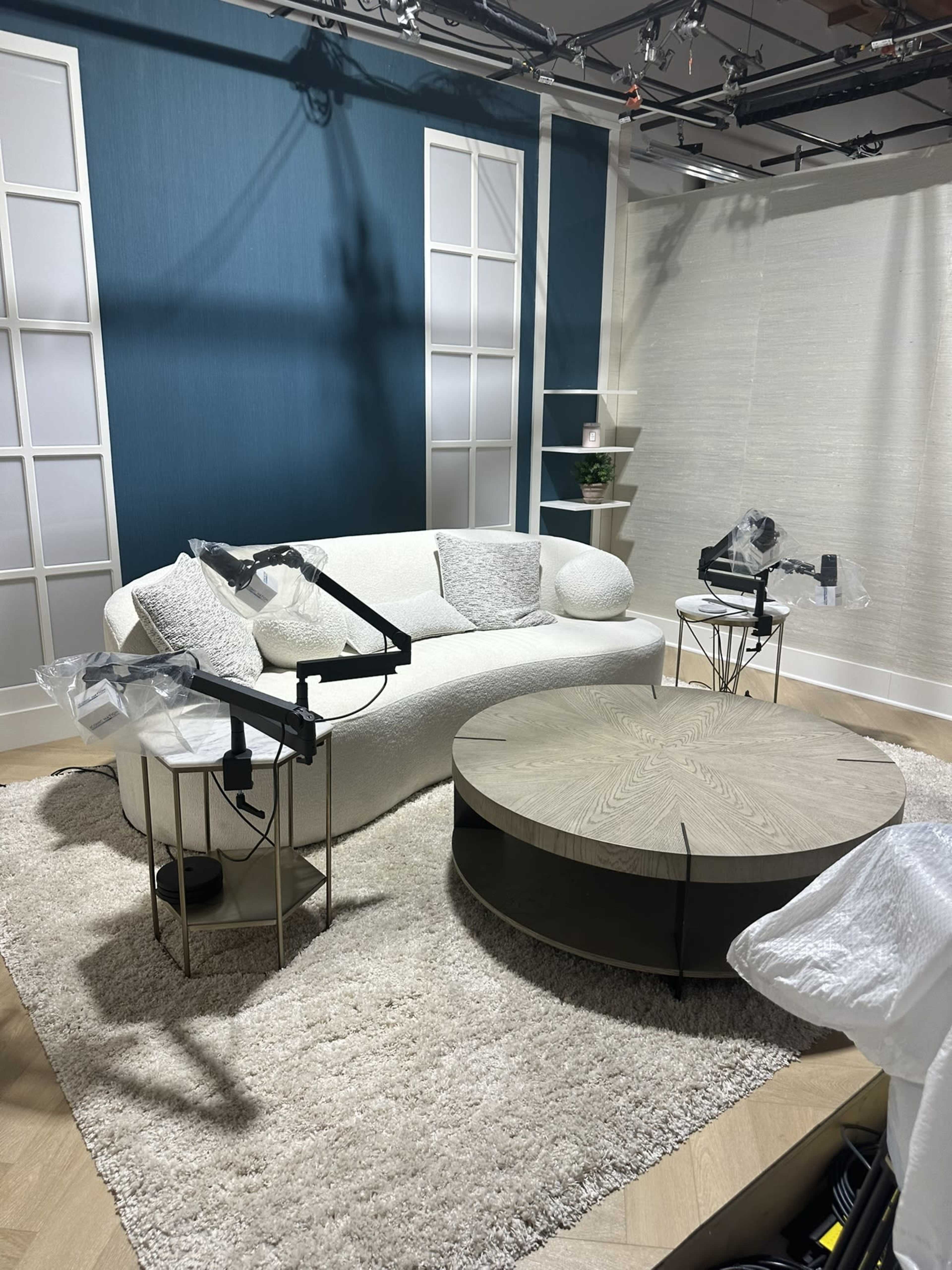 The image shows a modern studio setting featuring a white curved sofa, a round wooden coffee table, and light-colored decor against a teal backdrop.