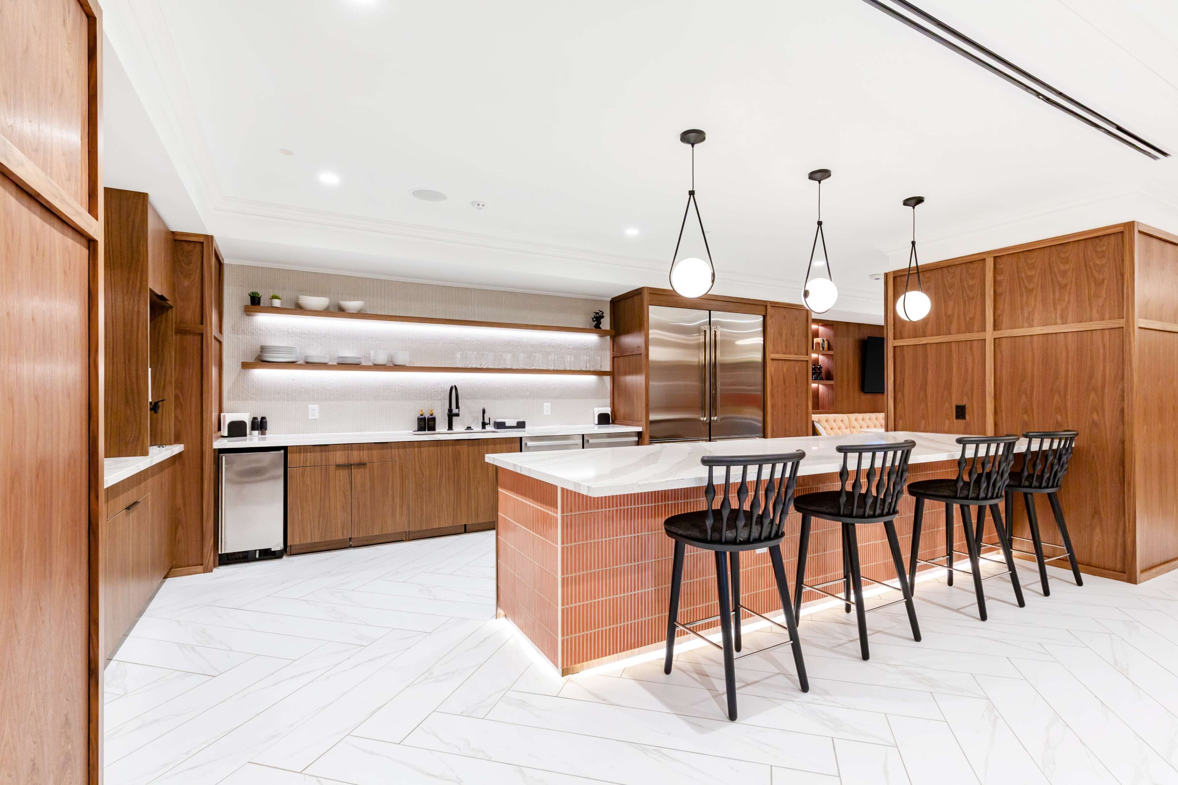 The image shows a modern kitchen with wooden cabinetry, a large island featuring a marble countertop, and black bar stools, illuminated by pendant lighting.