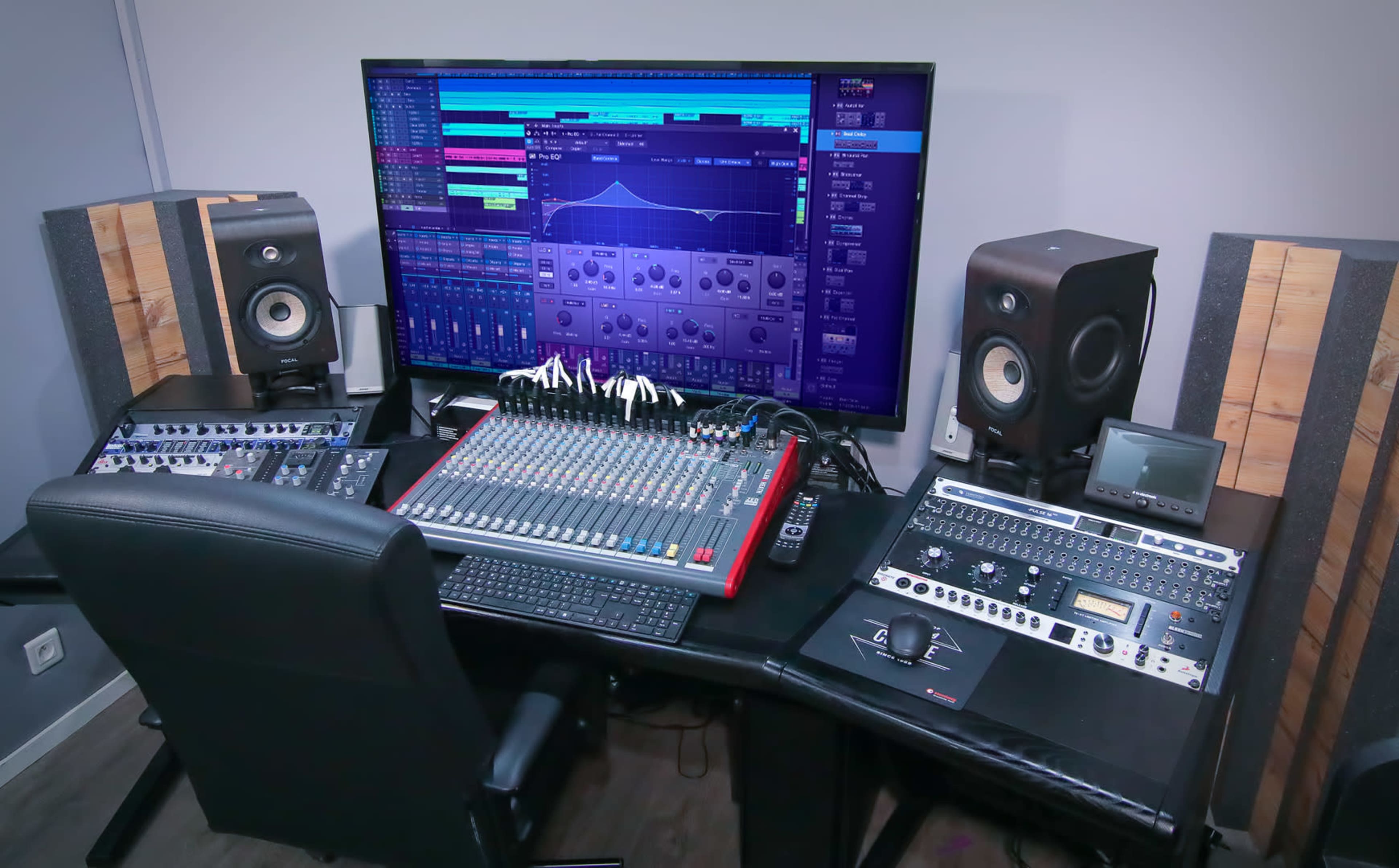 A home recording studio featuring a large monitor, a mixing console, audio speakers, and various audio equipment arranged on a desk.