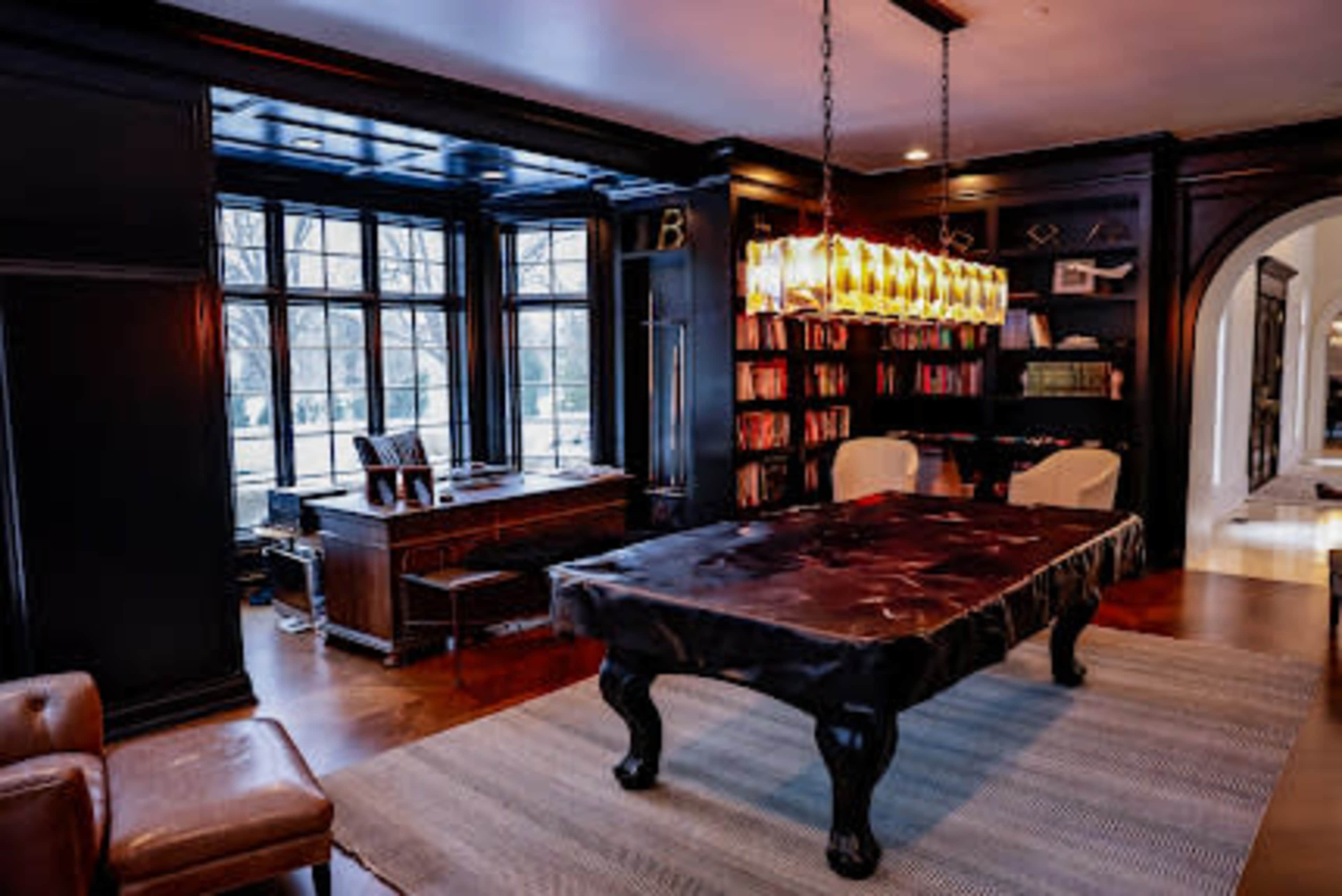 The image shows a well-furnished study or office space featuring a large wooden desk, a table with a dark surface, and a bookshelf filled with books, illuminated by pendant lighting.
