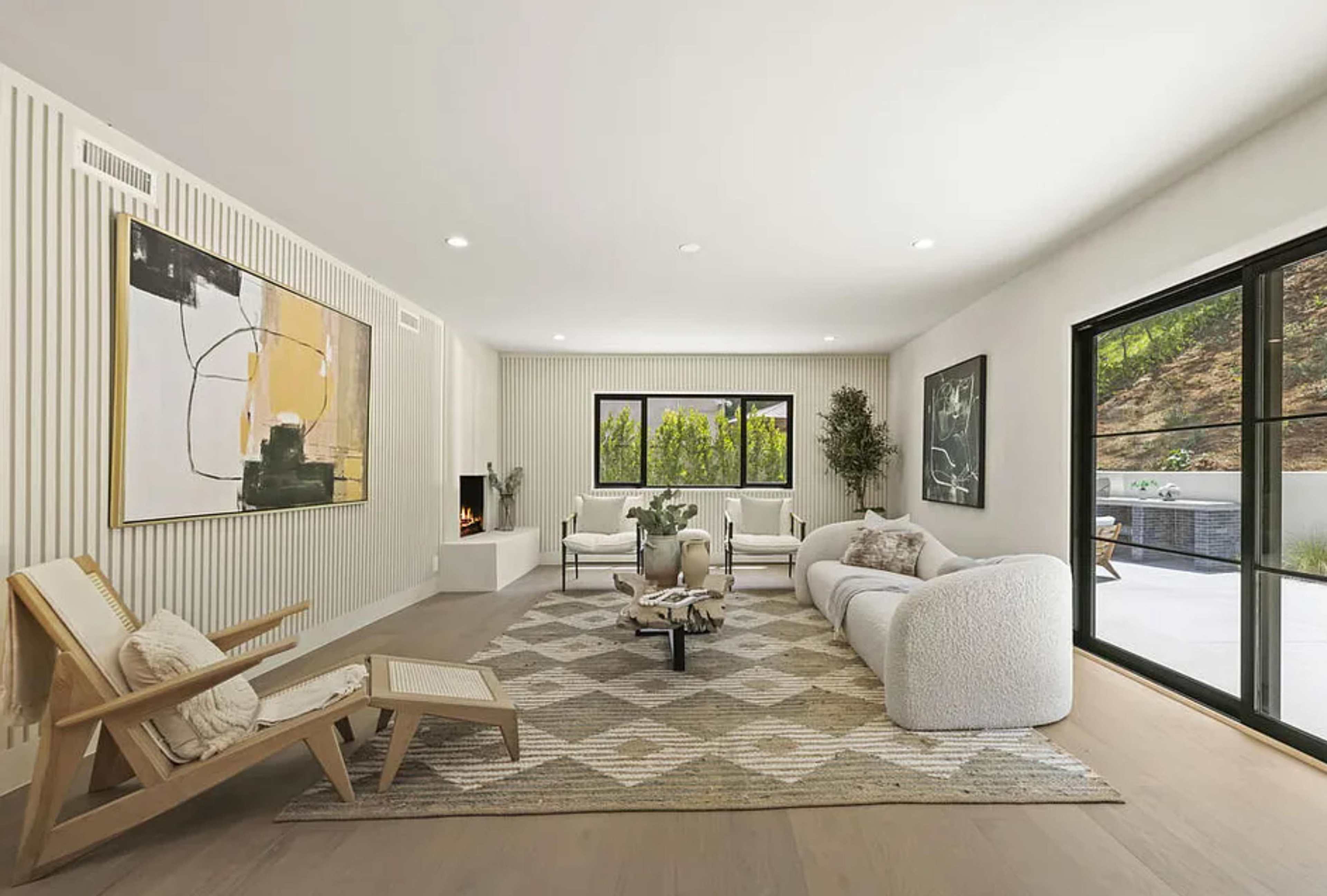 The image shows a modern living room featuring a light-colored sofa, an armchair, and a geometric area rug, with large windows providing a view of an outdoor space.