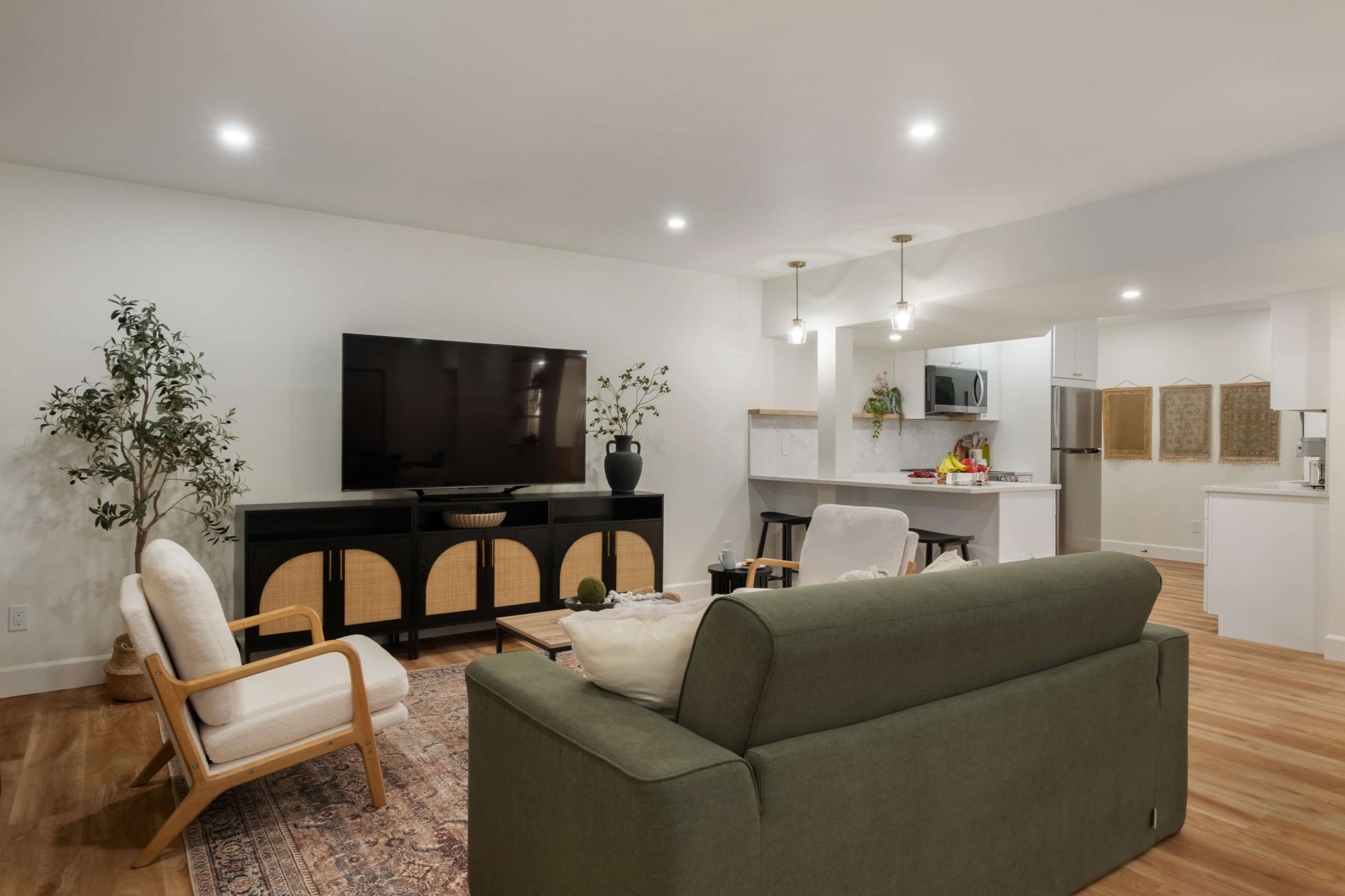 The image shows a modern living room with a green sofa, a television mounted on the wall, and a kitchen area visible in the background.