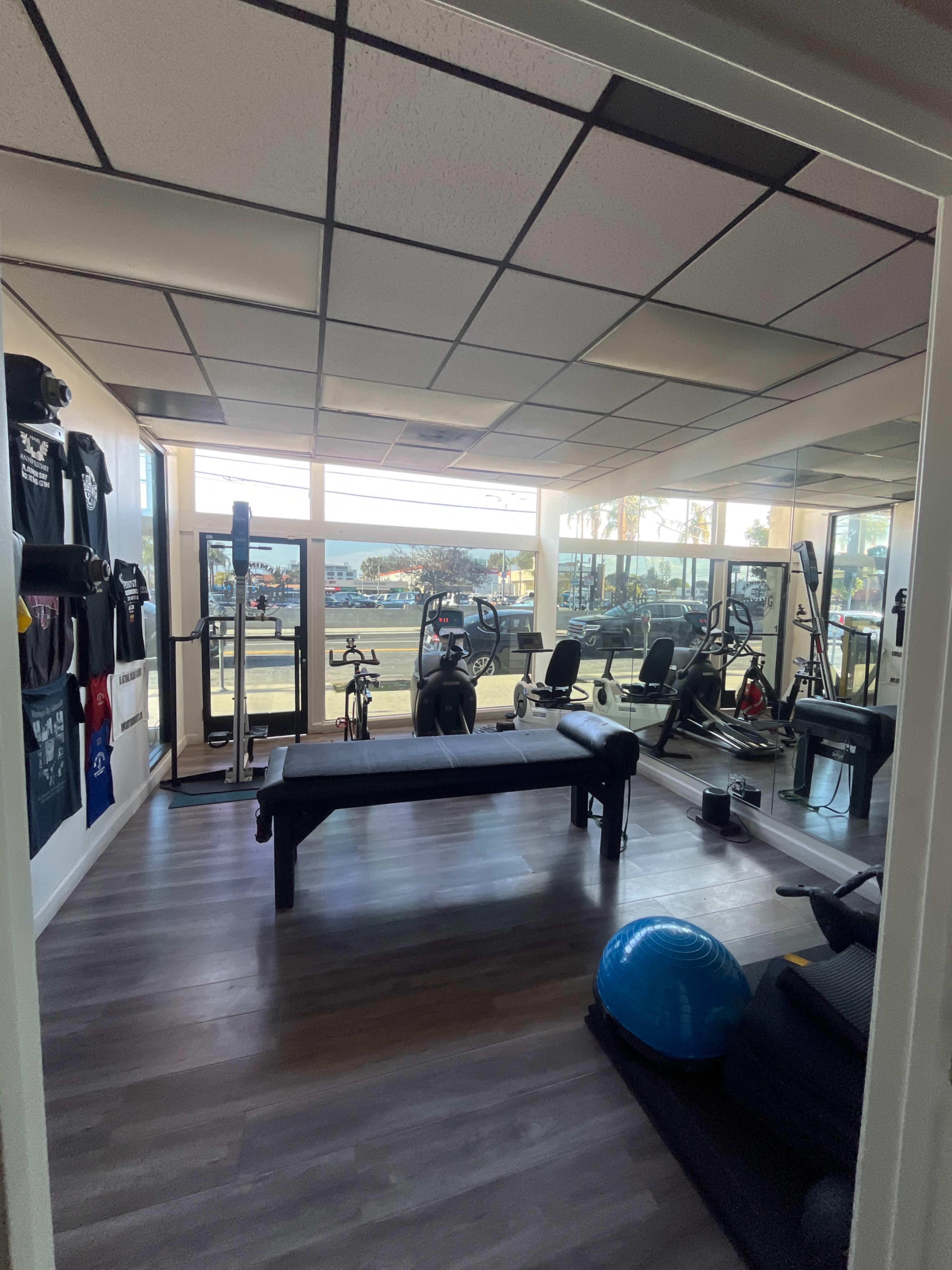 The image shows a fitness room with gym equipment, a bench, and large windows allowing natural light.