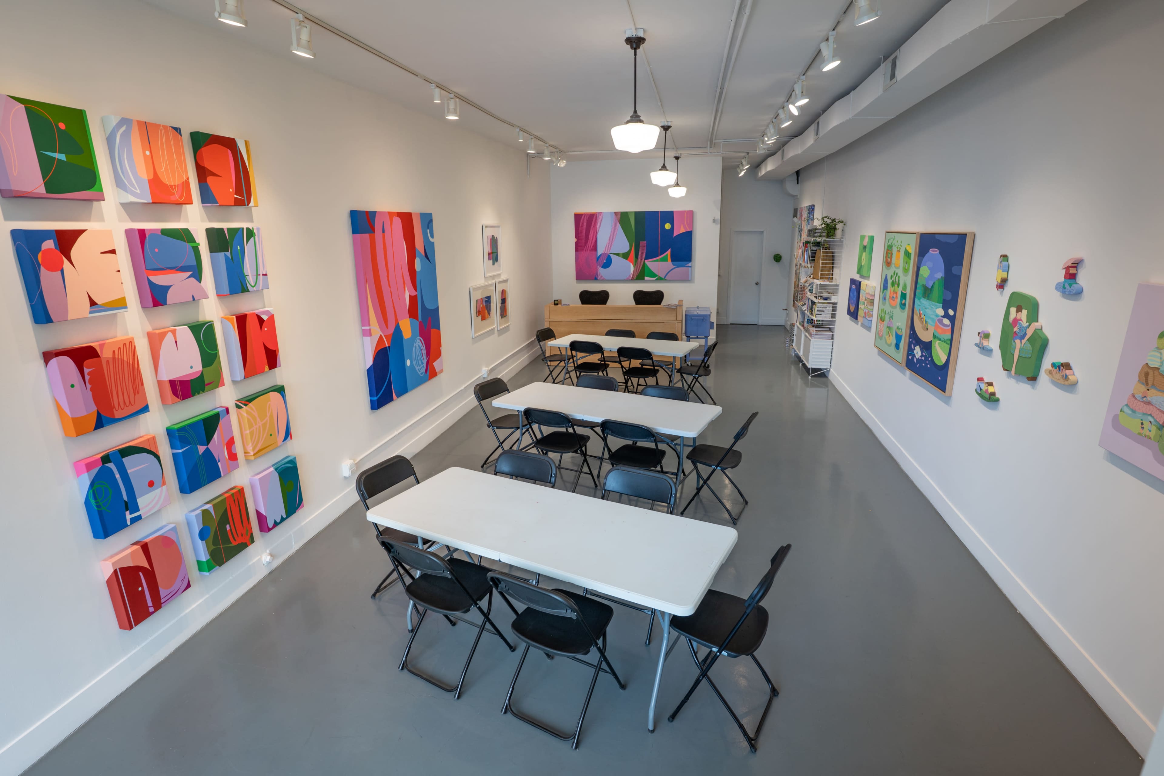 The image shows an art gallery with colorful abstract paintings on the walls and rows of black folding chairs and rectangular tables arranged in the center.