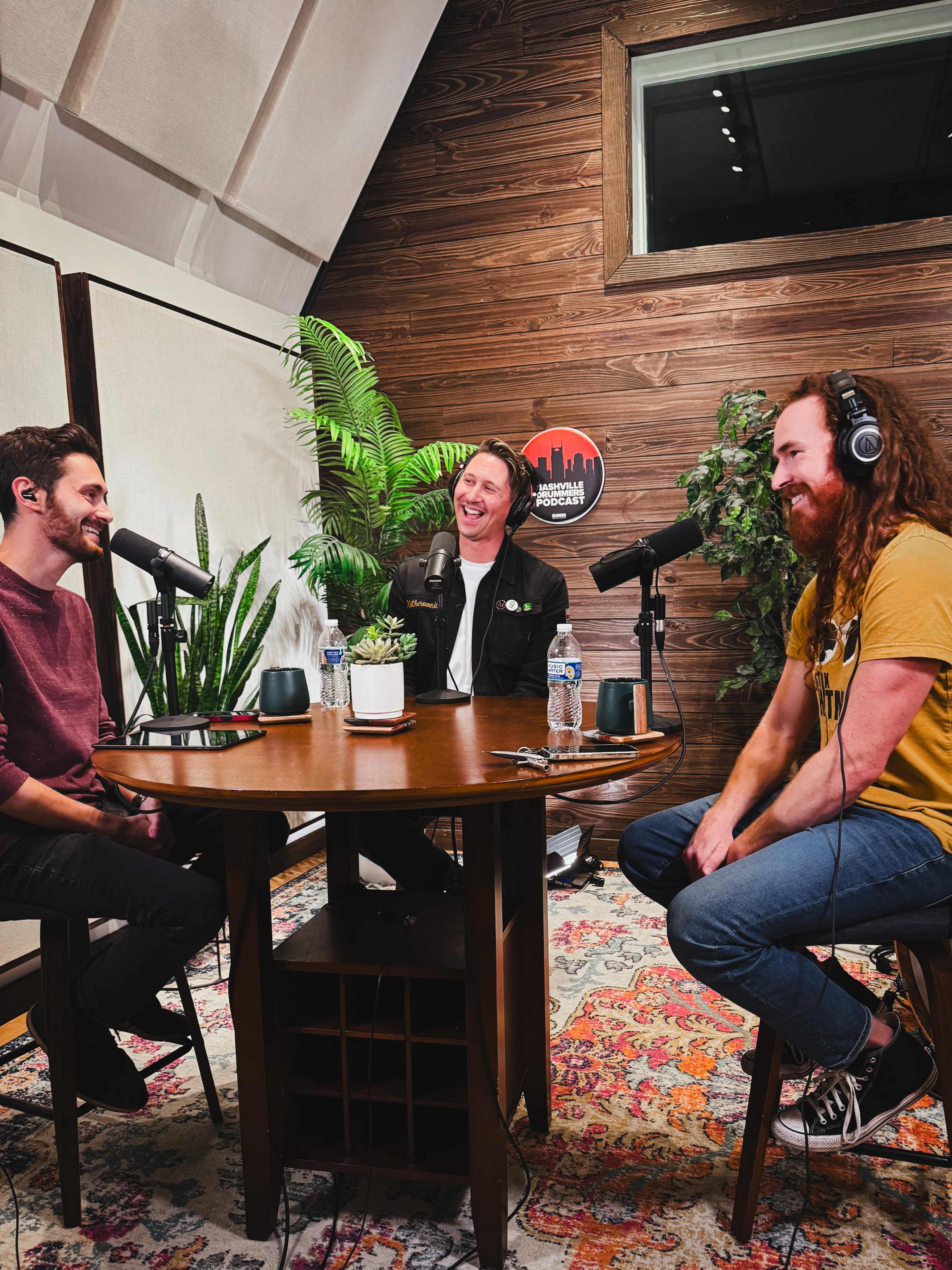 Three men sit around a wooden table in a recording studio, each wearing headphones and speaking into microphones, with potted plants in the background.