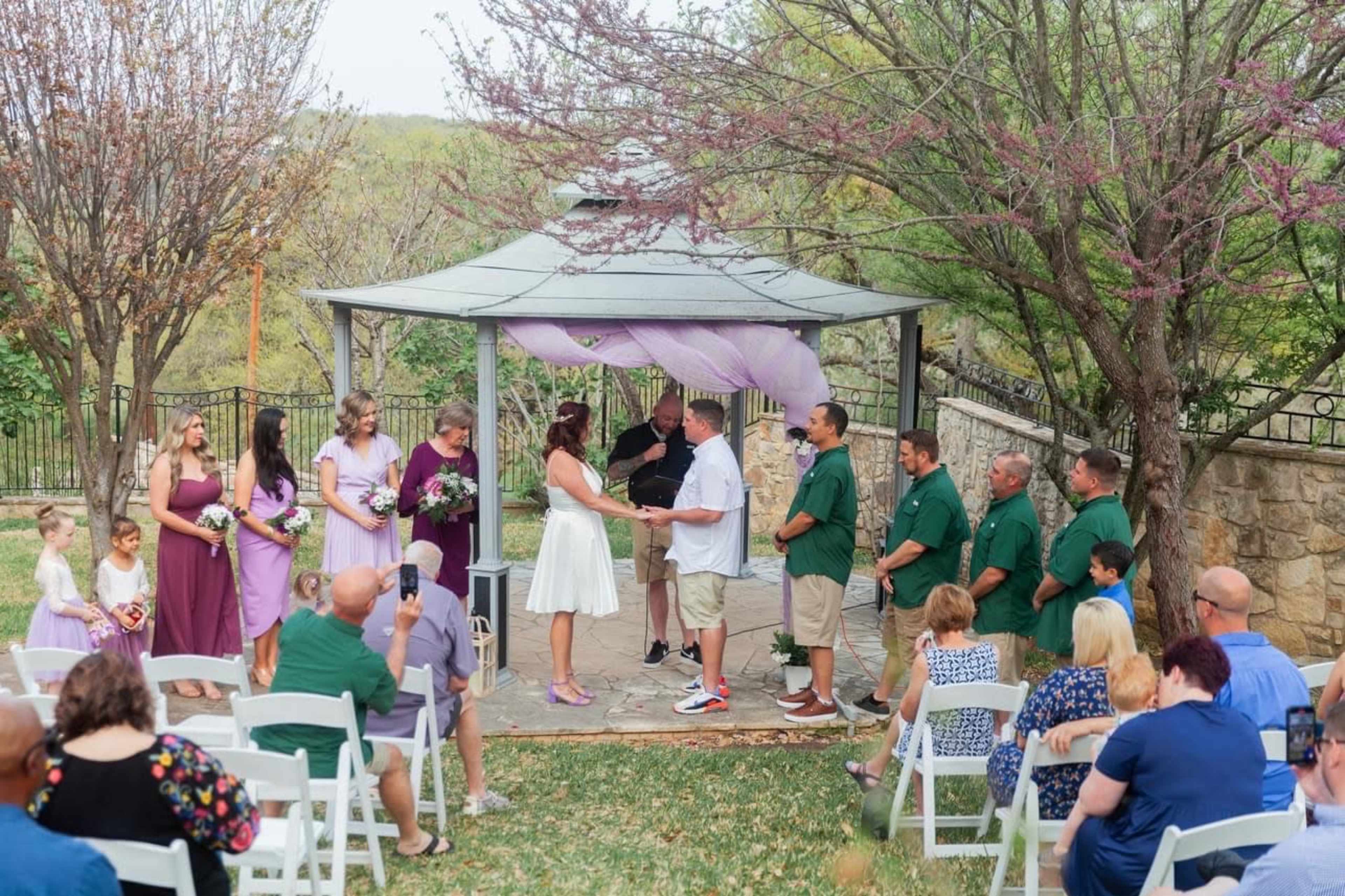 A couple exchanges vows under a gazebo surrounded by wedding guests and floral decorations.