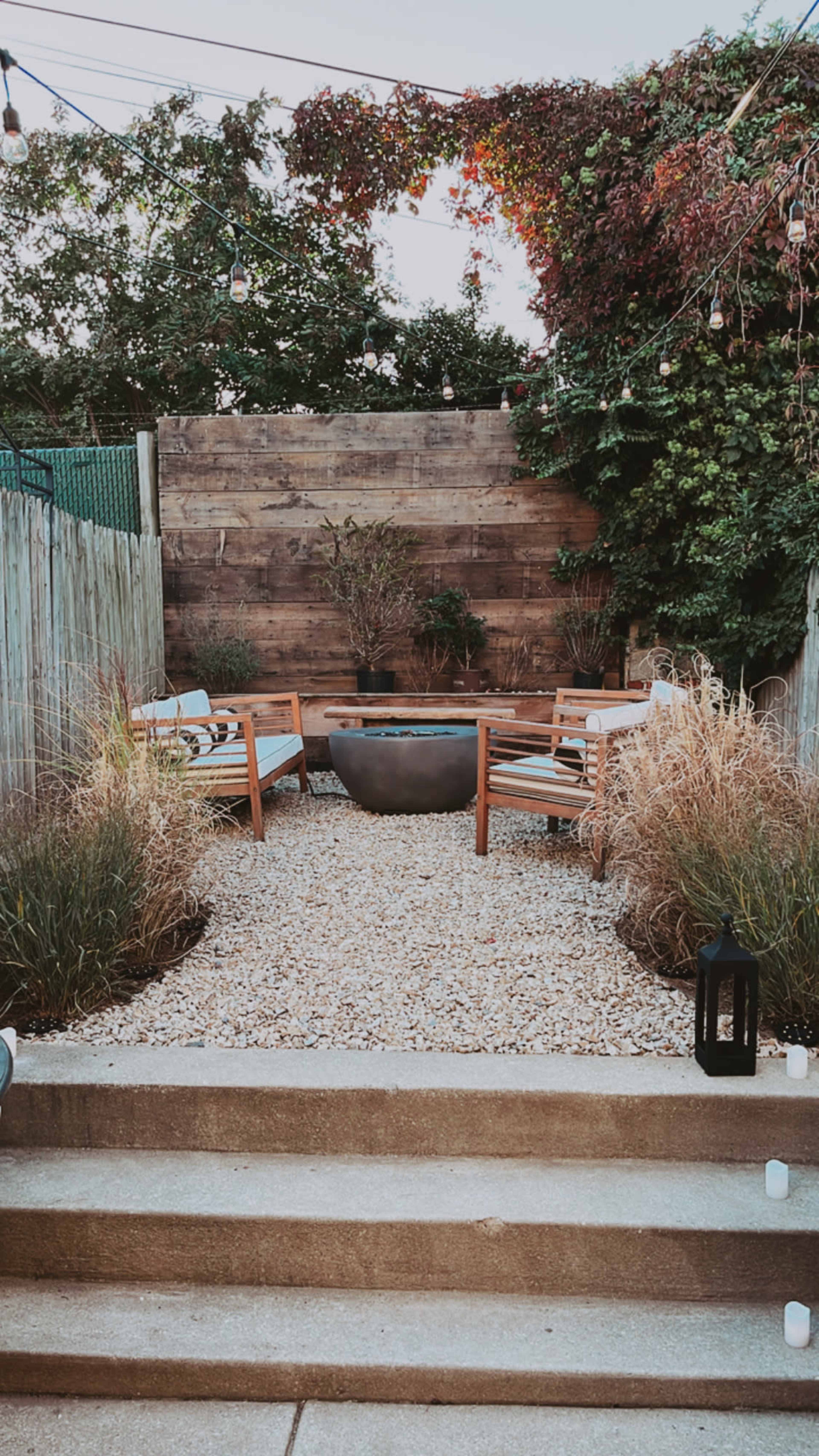 A gravel patio features a circular fire pit surrounded by wooden seating and lush greenery in a fenced garden.