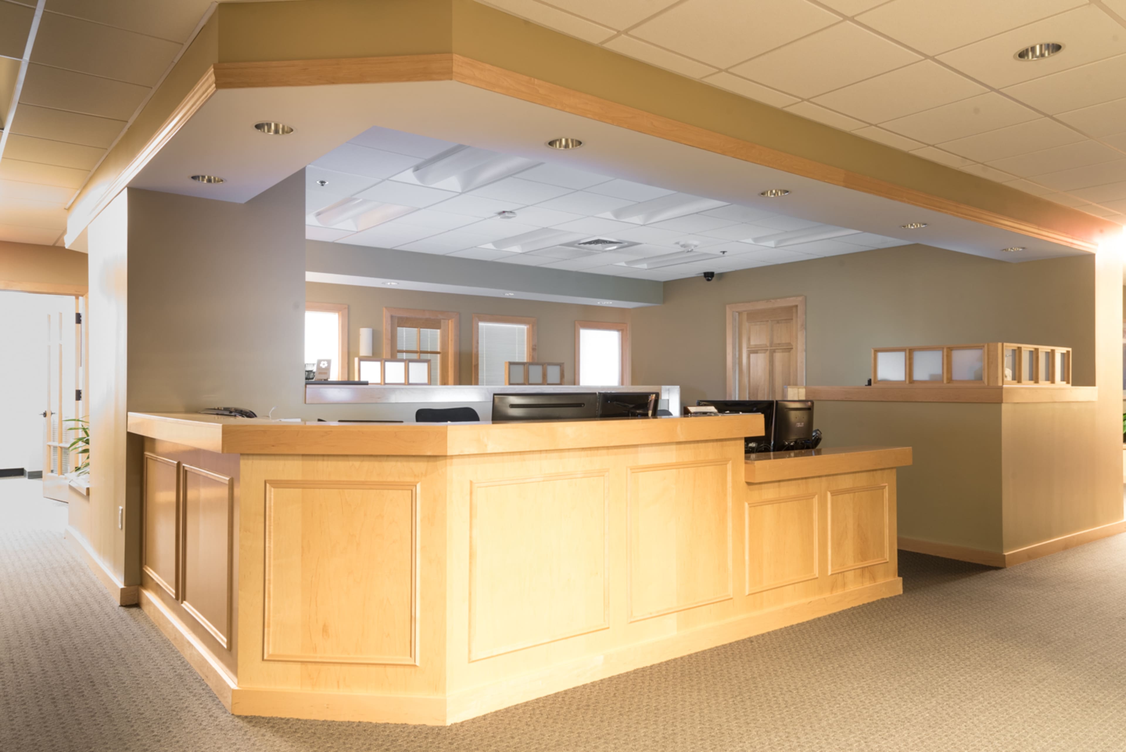 A spacious office reception area featuring a wooden front desk with multiple workstations and light-colored walls.